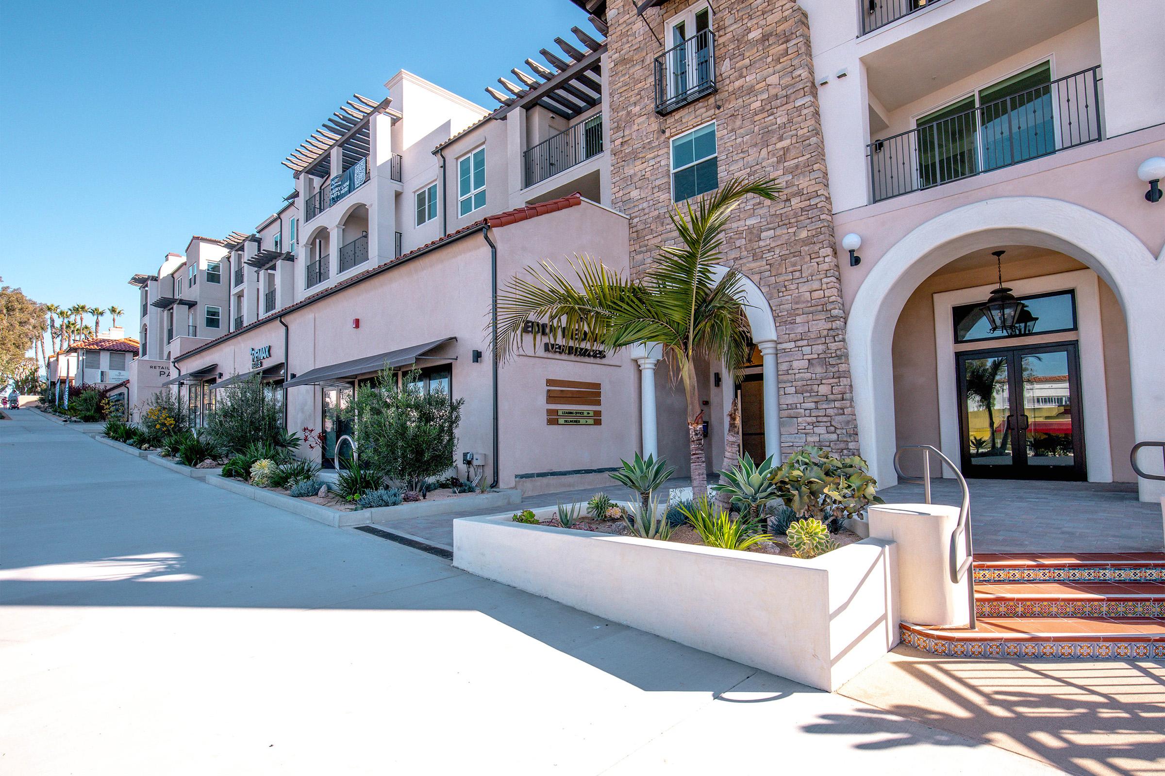 A modern multi-story building with a mix of architectural styles, featuring stucco and stone facades. The entrance has a welcoming staircase and decorative tiling, surrounded by greenery and planters. The street is lined with shops and restaurants, under a clear blue sky.