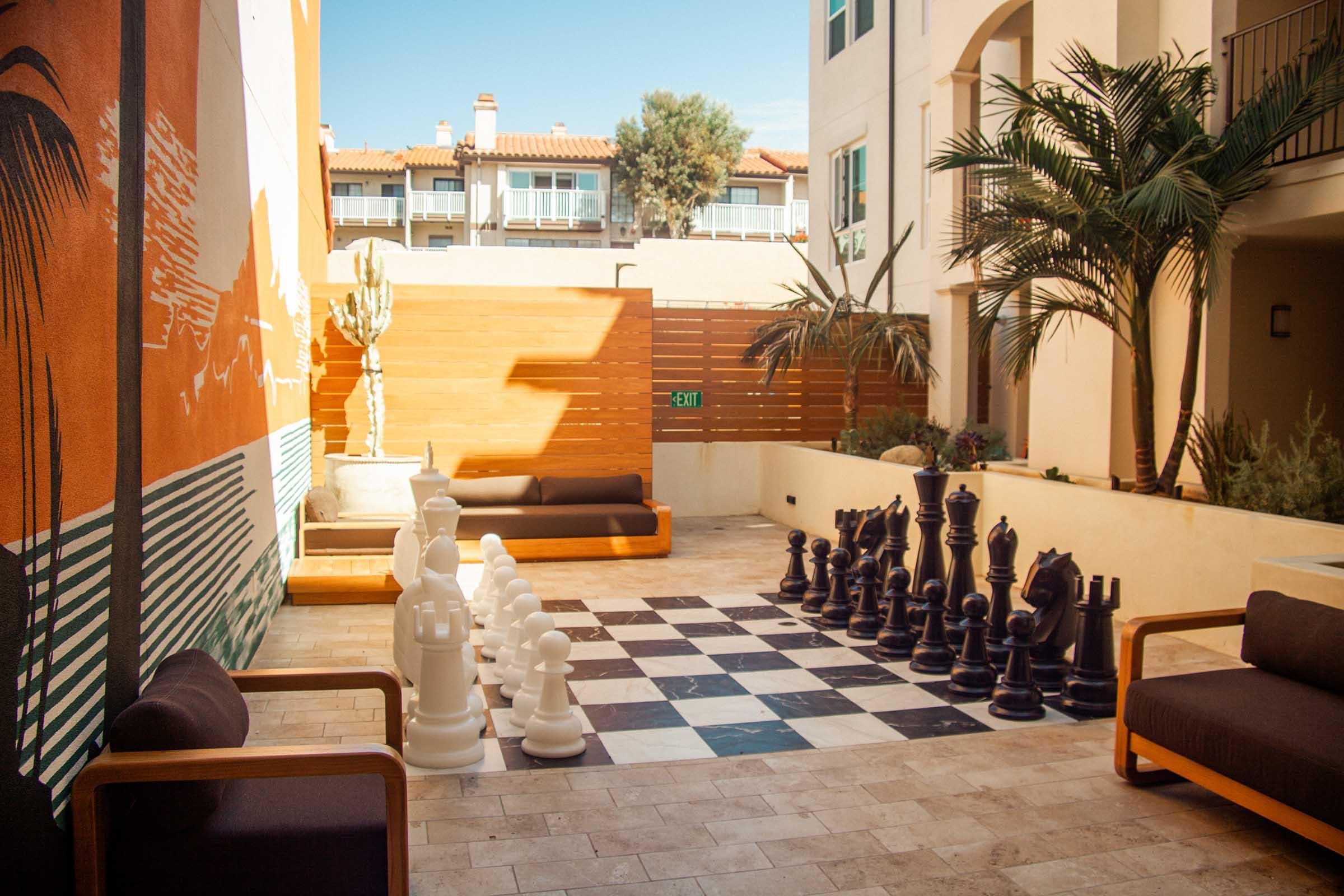 A spacious courtyard featuring a large outdoor chess set with white and black pieces on a checkered board. Surrounding the chess area are modern seating options and greenery, including palm trees and a decorative wall mural, creating a vibrant and inviting atmosphere.
