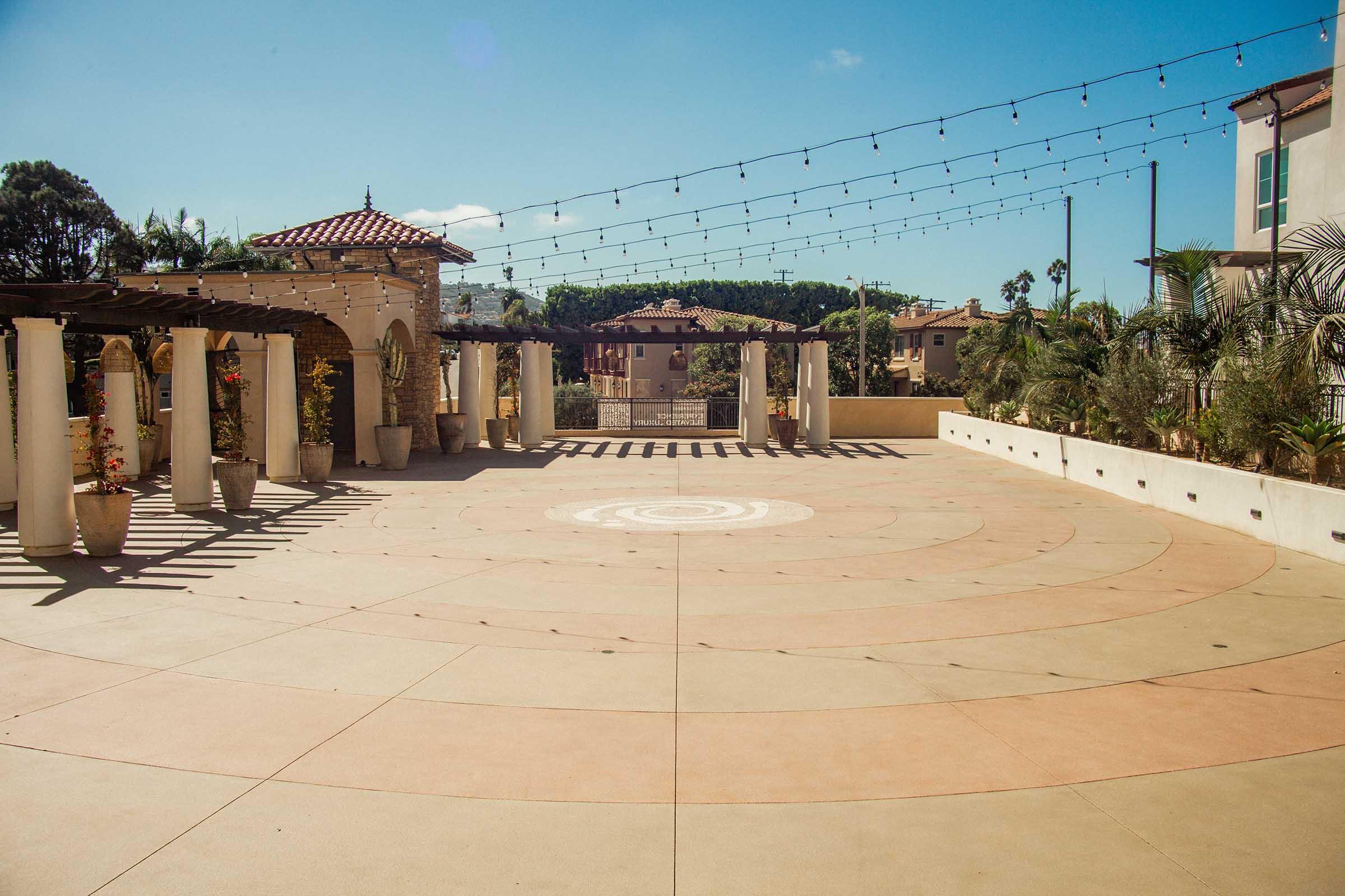 A spacious outdoor area featuring a circular pattern on the ground, surrounded by pillars and potted plants. String lights hang overhead, creating a welcoming atmosphere. In the background, a glimpse of buildings and palm trees can be seen under a clear blue sky.