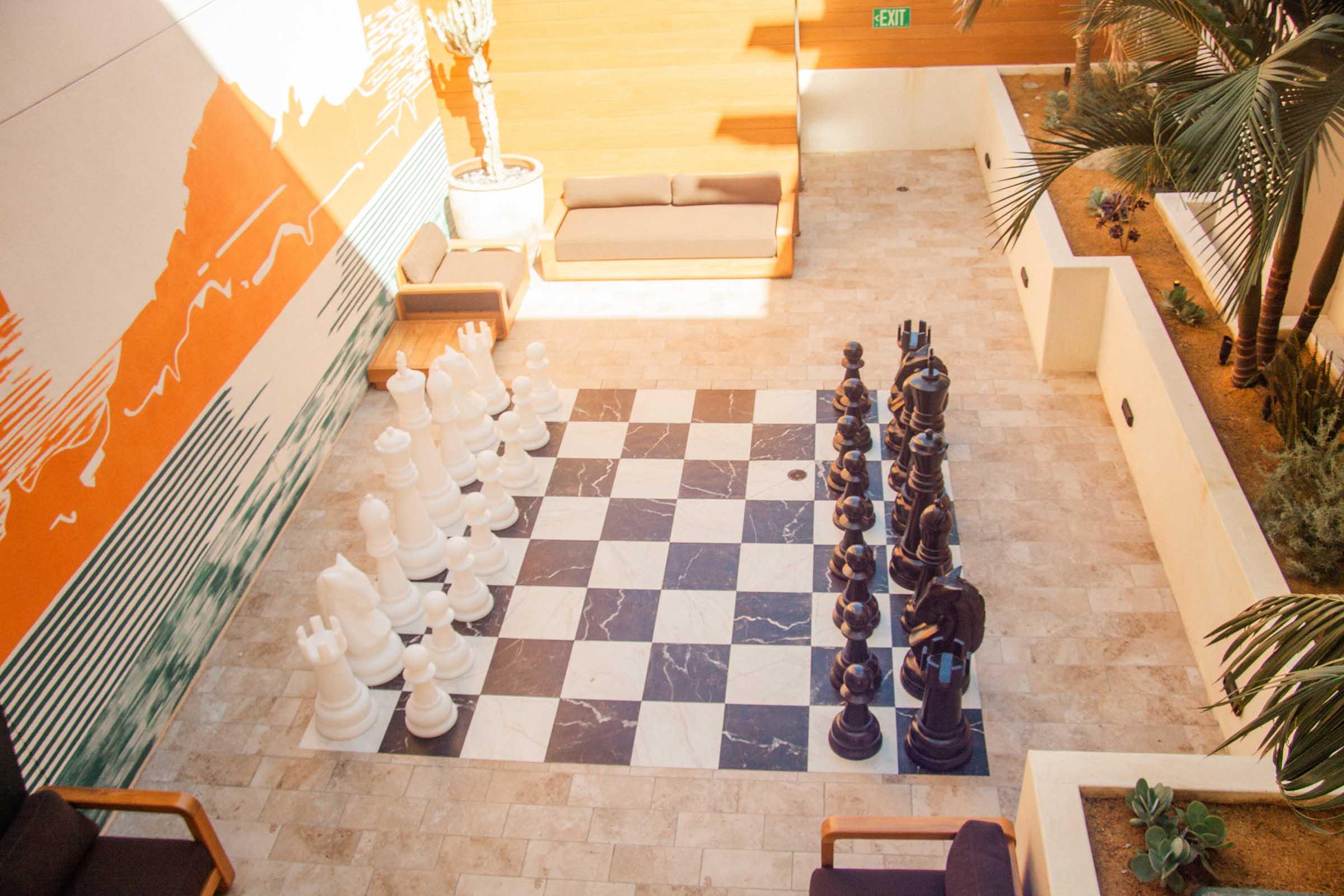Large outdoor chessboard with oversized black and white pieces, set in a spacious area decorated with modern furnishings and plants. Natural light illuminates the scene, highlighting the contrasting colors of the chess pieces and the stylized mural on the wall. Comfortable seating is visible in the background.