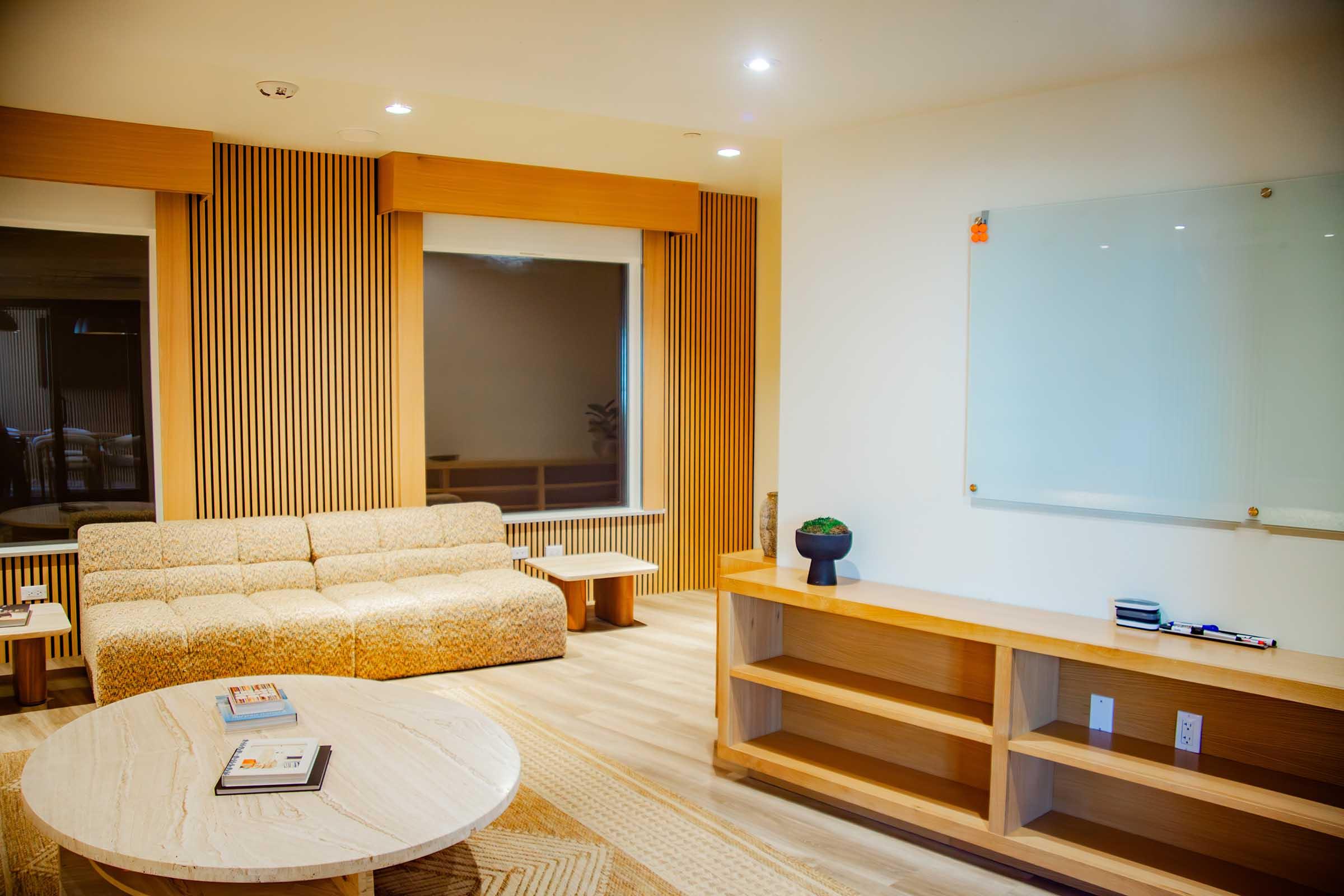 A modern, well-lit living space featuring a beige sofa, a round coffee table, and a wooden media console. The walls are adorned with vertical wooden panels, and there’s a whiteboard mounted on the wall. Two large windows allow natural light to fill the room.