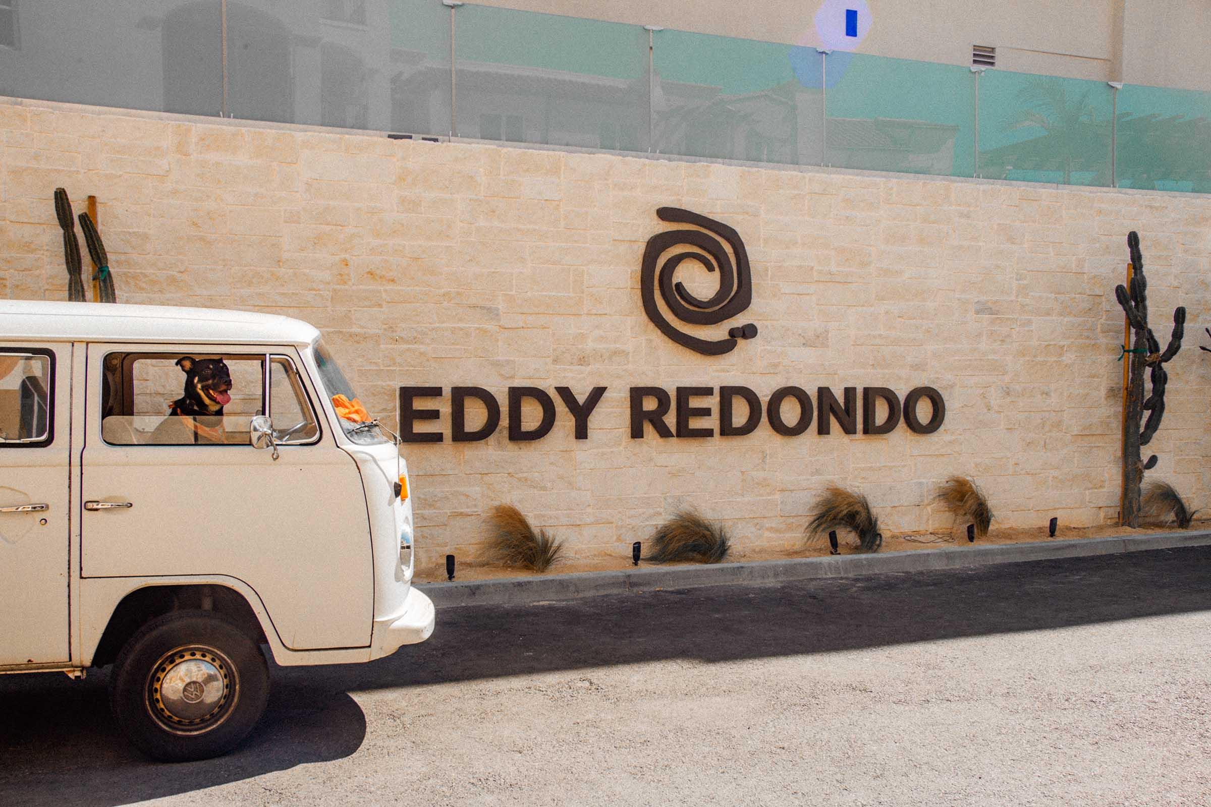 A vintage white van is parked next to a stone wall featuring the text "EDDY REDONDO." The wall is accented with decorative cacti and desert plants, creating a warm, inviting atmosphere. The scene suggests a blend of retro charm and modern design, ideal for a welcoming establishment.