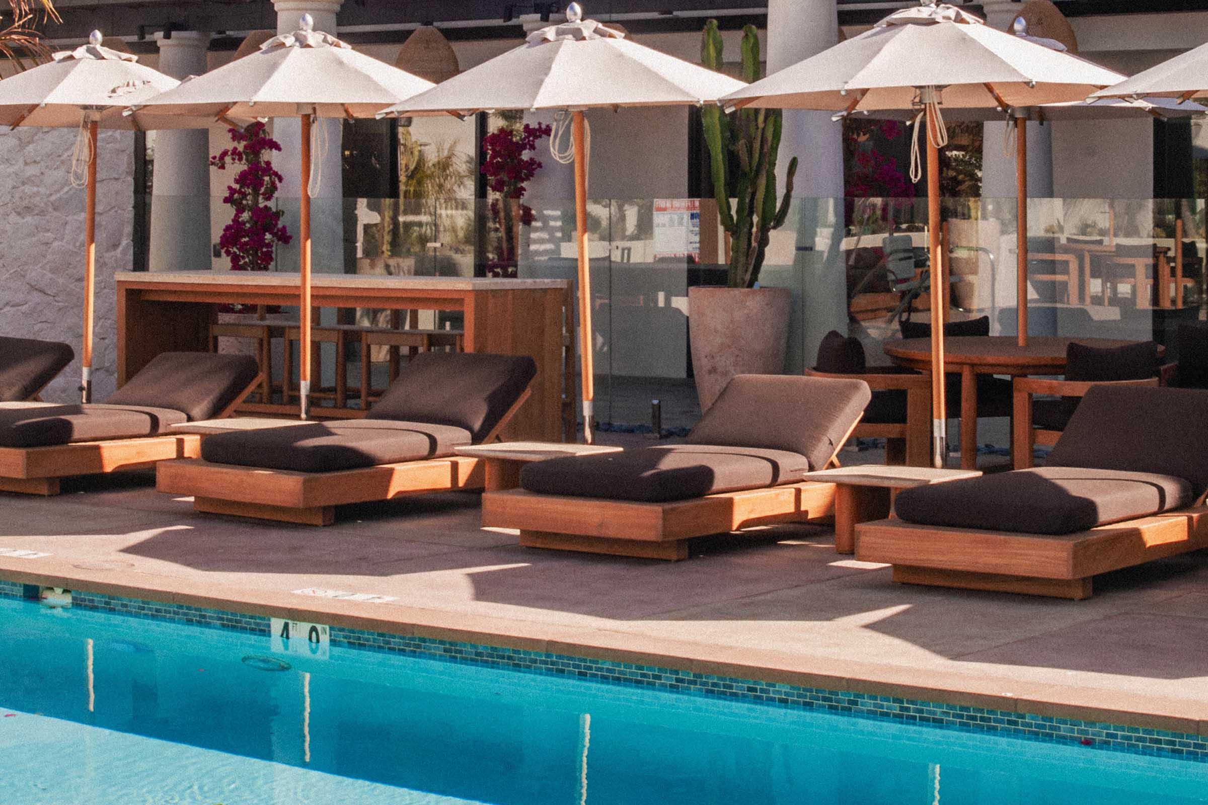 Lounge chairs with dark cushions are arranged next to a swimming pool, under large umbrellas. A wooden bar area is visible in the background, surrounded by plants and decorative elements, creating a relaxing outdoor environment.