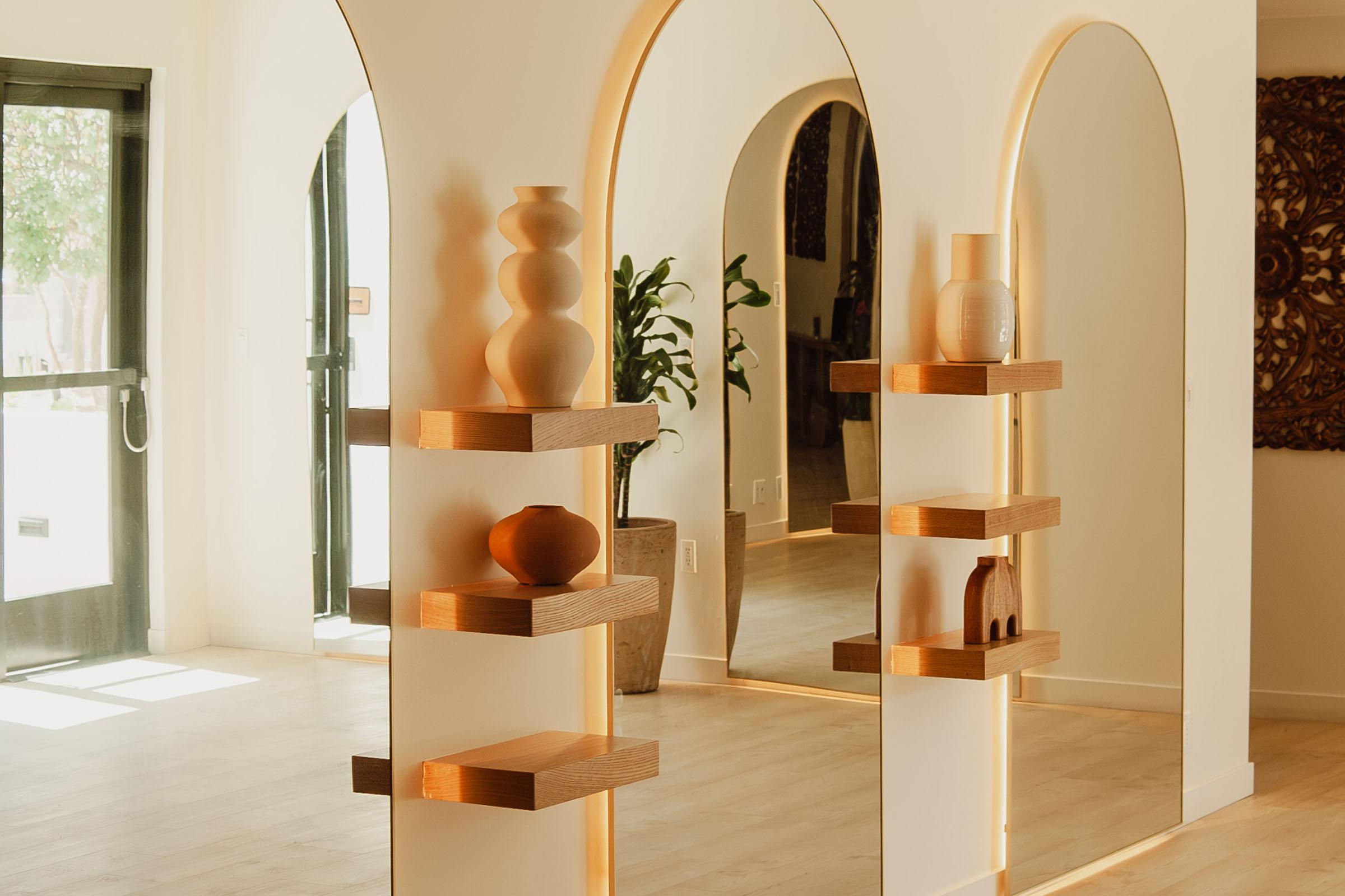 A modern interior scene featuring a decorative shelving unit with various vases displayed. The shelves are illuminated, creating a warm ambiance. In the background, a large arch mirrors the shelves and a potted plant, with an entrance visible on the left. The overall design is minimalistic and stylish.