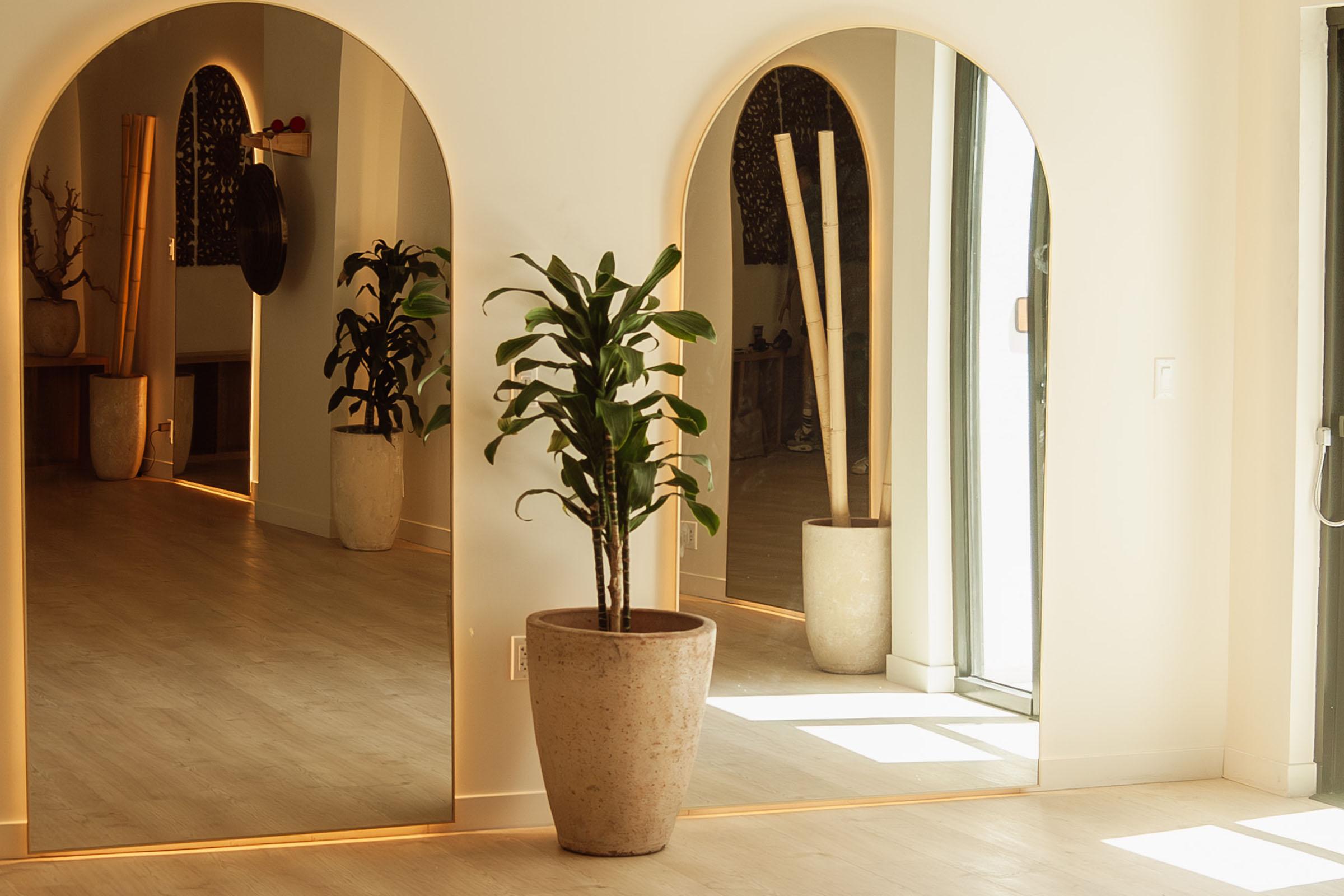 A modern interior featuring three arched mirrors on a light-colored wall. In the foreground, a potted plant adds greenery, while warm light enhances the serene atmosphere. The floor is light wood, and the space reflects a minimalist aesthetic with elegant decor elements in the background.