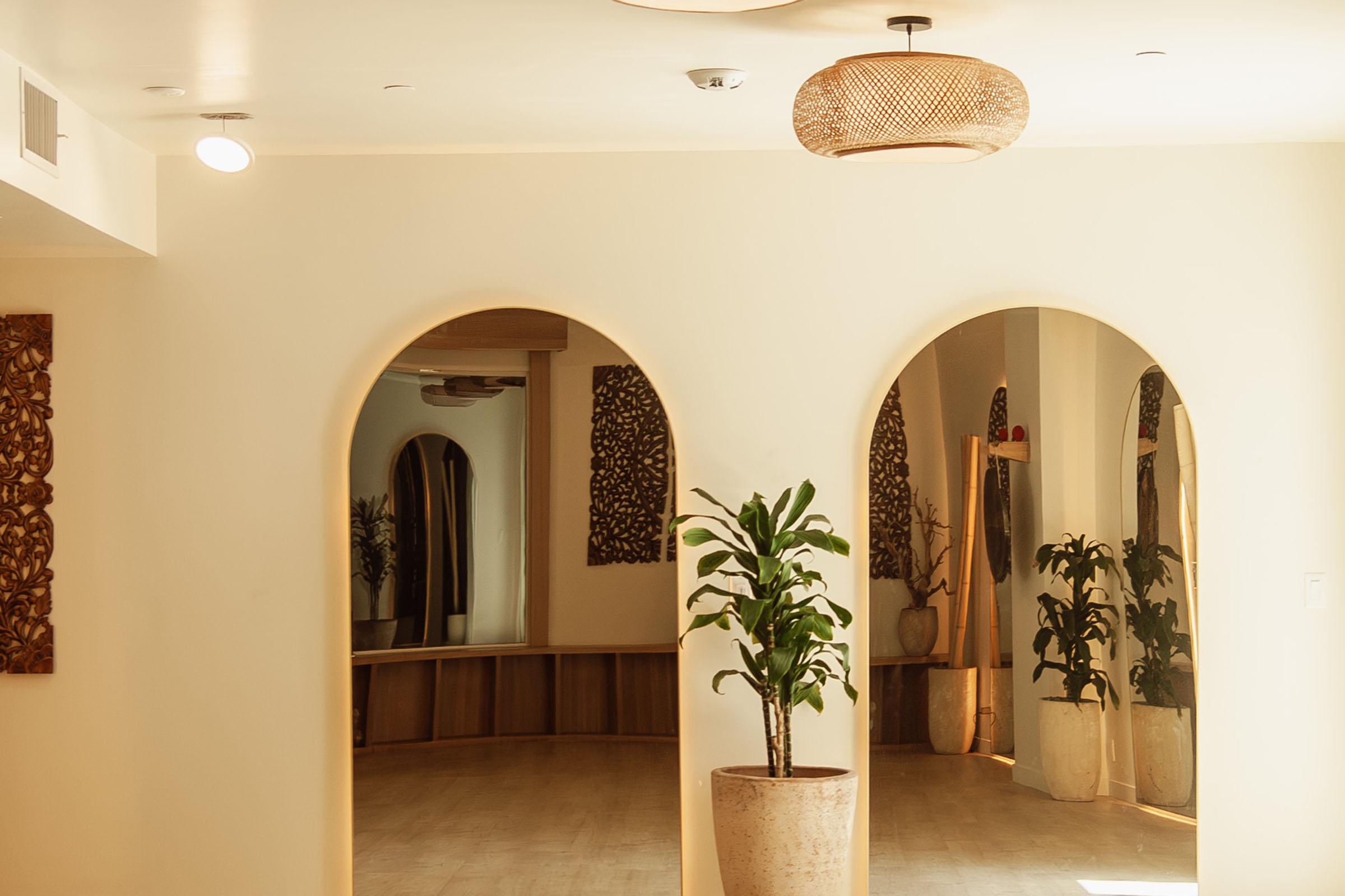 A bright, minimalist interior space featuring three arched doorways. Two potted plants are visible, one in the foreground and another in the background. The walls are a soft cream color, and a woven pendant light hangs from the ceiling. Decorative elements include wood textures and subtle patterns.