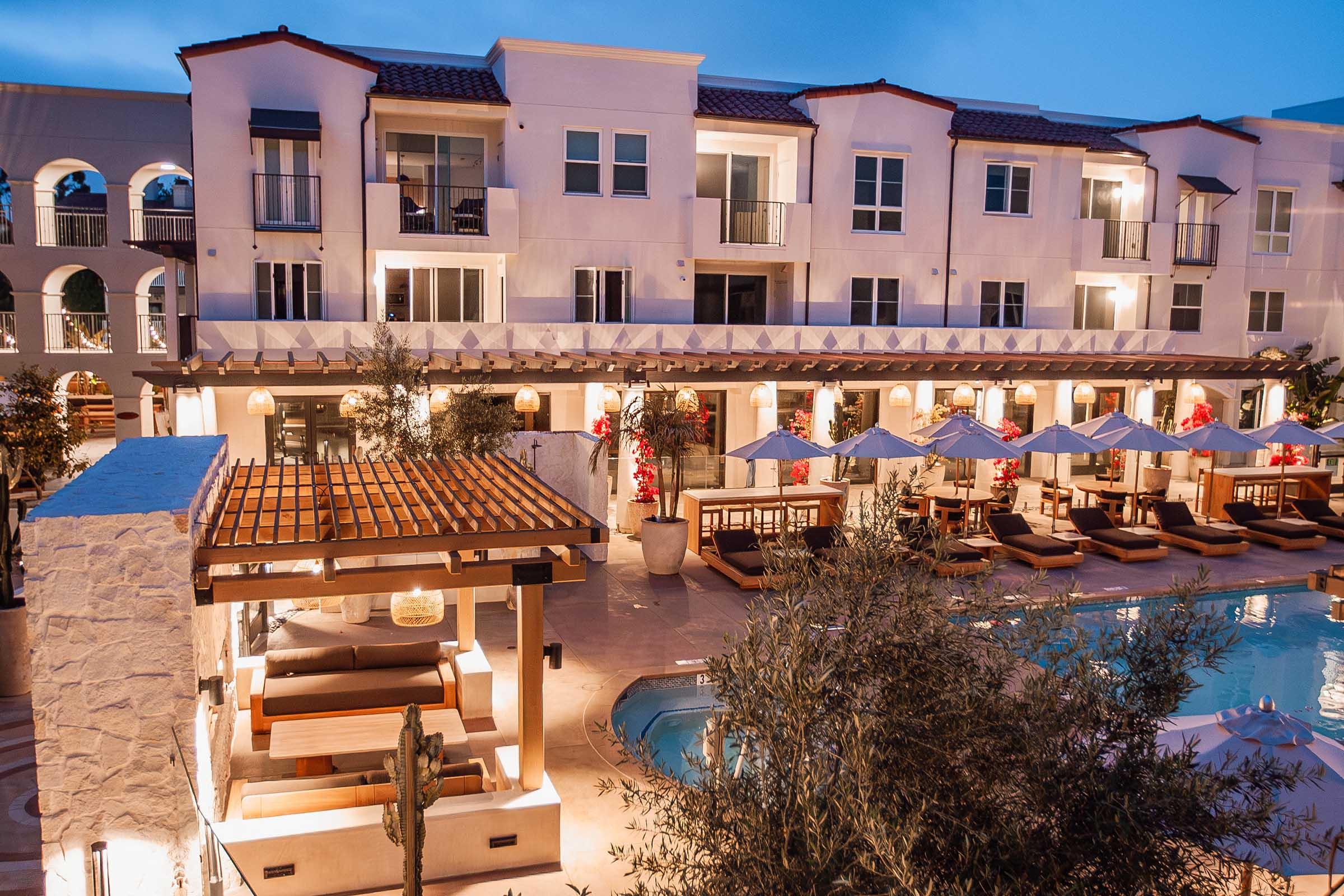 A luxurious hotel courtyard featuring a elegantly designed pool area surrounded by lounge chairs and umbrellas. The building has multiple balconies and white stucco walls, with soft lighting creating a warm ambiance in the evening. Olive trees and a wooden pergola enhance the aesthetic appeal of the space.