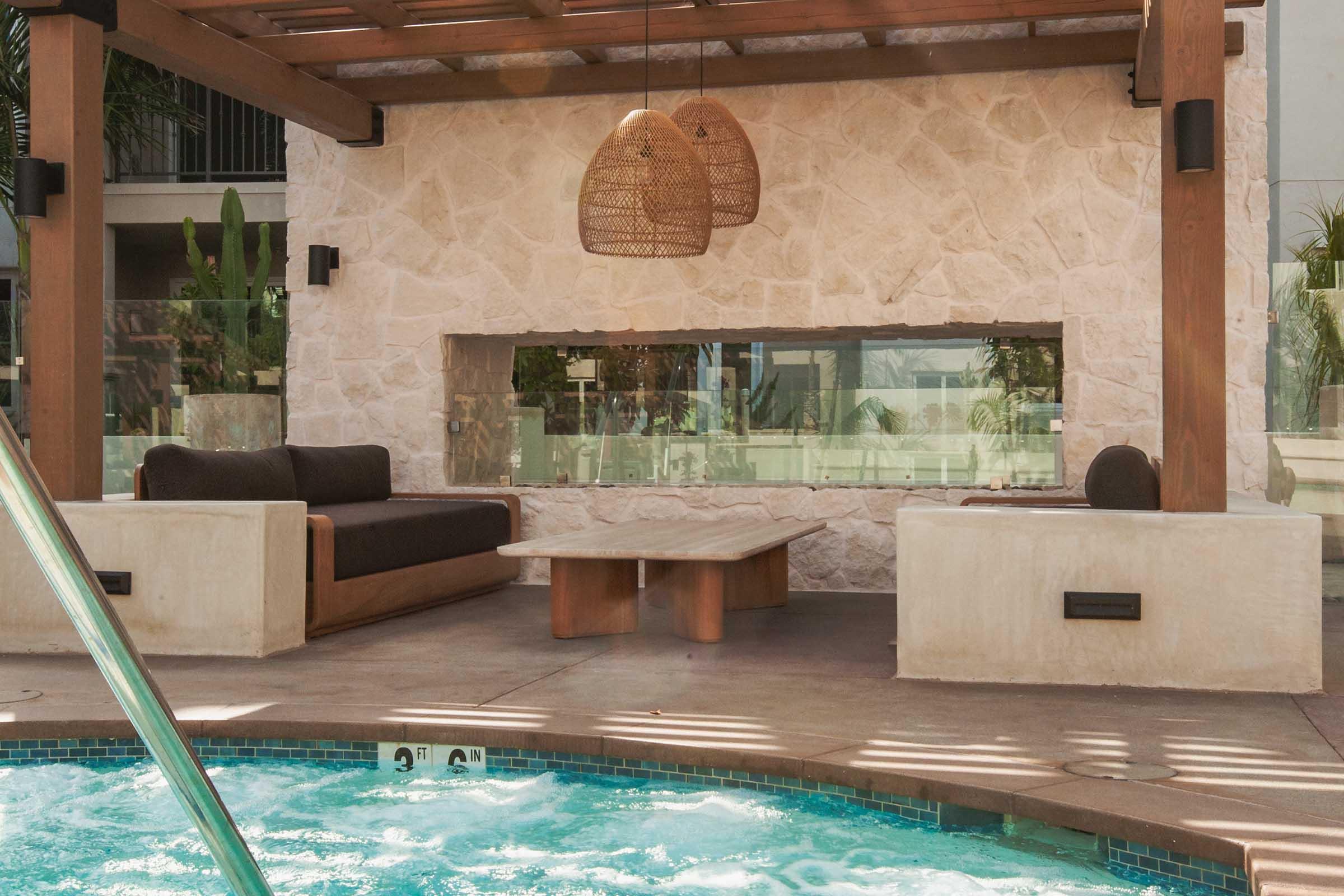 A stylish outdoor lounge area featuring a modern stone wall, a wooden table, and comfortable seating. Elegant wicker pendant lights hang above, and a pool with blue water is visible in the foreground. Lush greenery adds a touch of nature to the relaxing atmosphere.