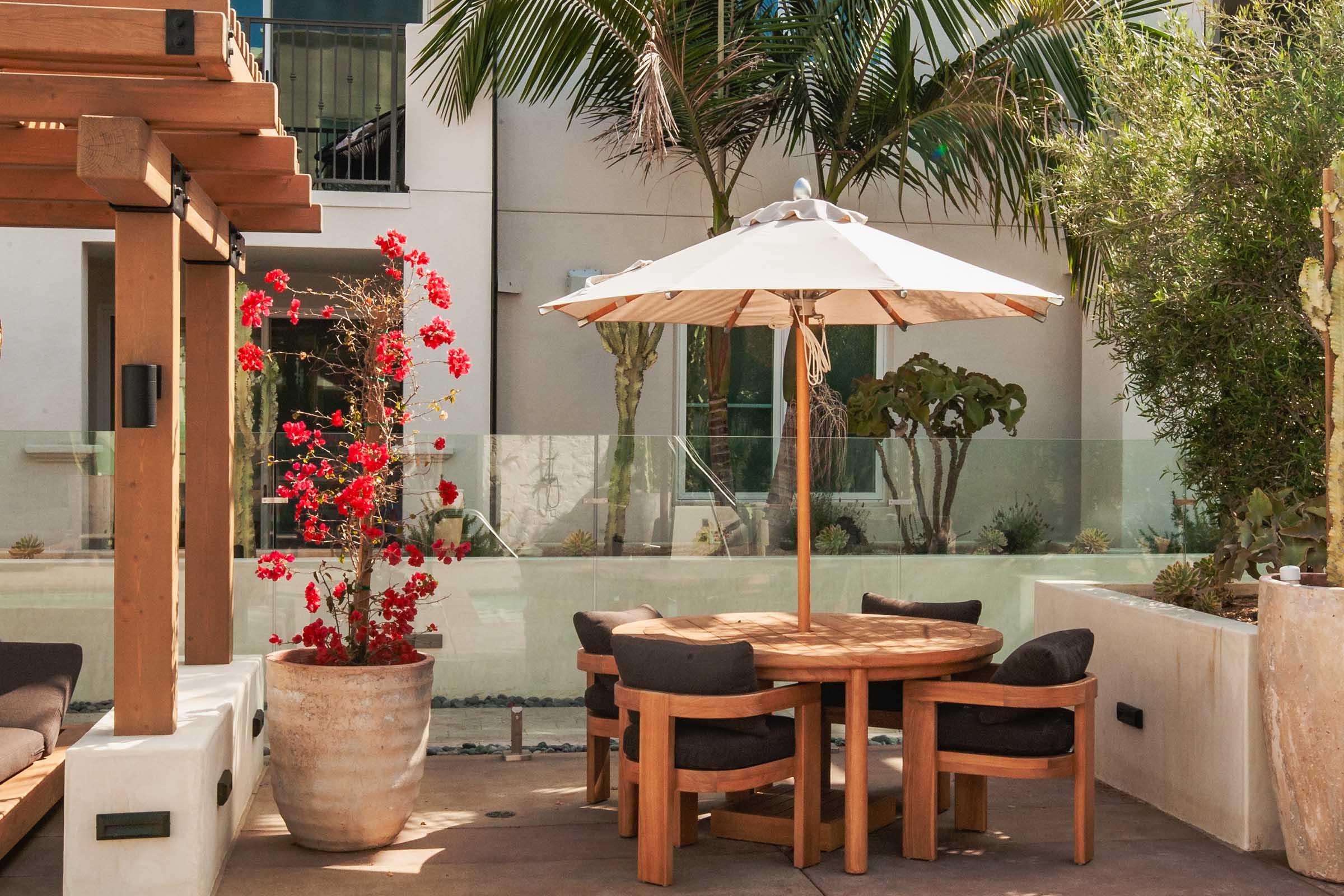 A sunny outdoor patio featuring a wooden table surrounded by brown chairs, an umbrella for shade, and a large potted plant with vibrant red flowers. The background includes a glass wall and tropical greenery, creating a relaxing and inviting atmosphere.