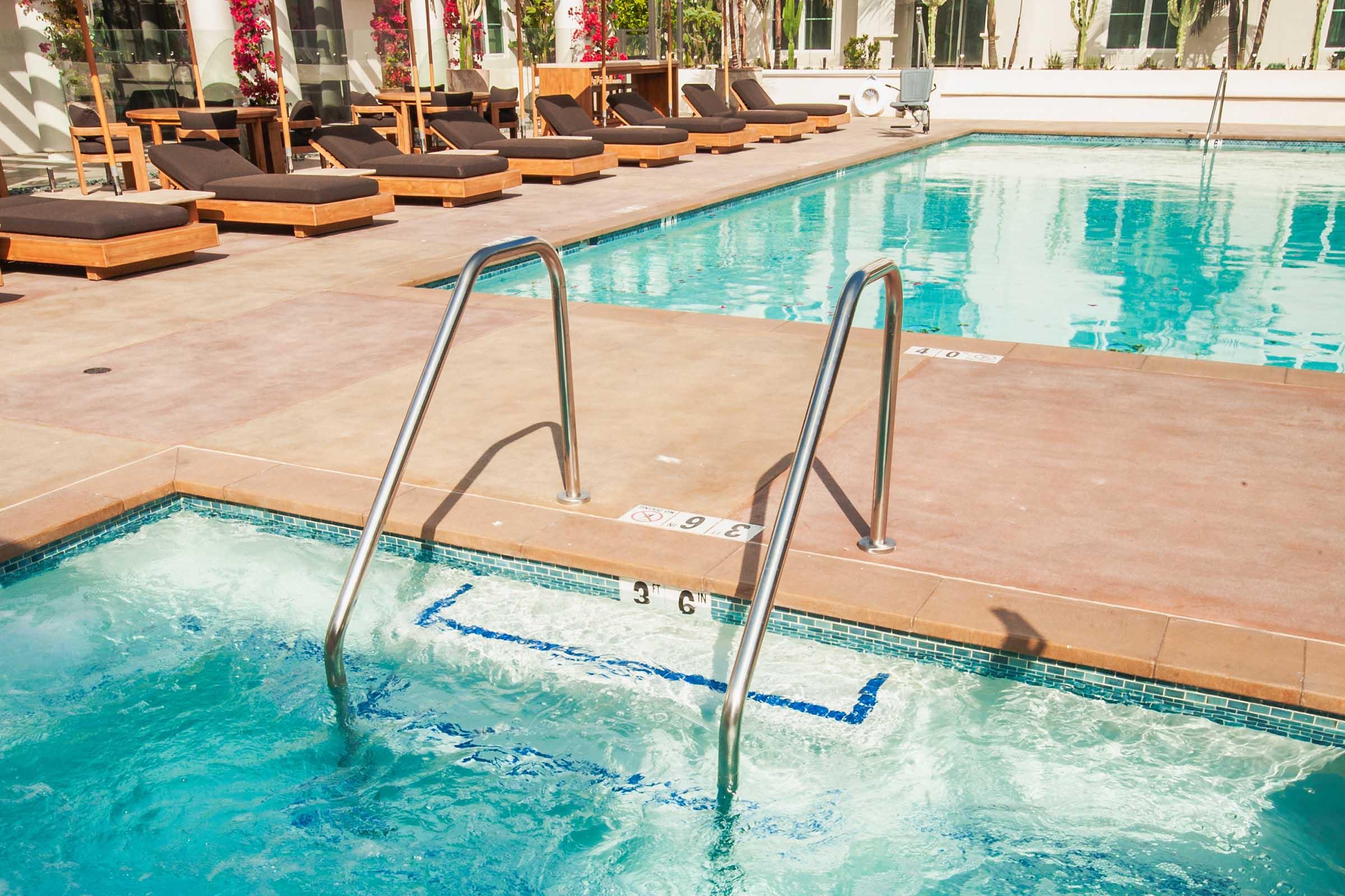 A clear, inviting swimming pool with a shallow area marked at 3 feet deep. Stainless steel steps lead into the water, surrounded by lounge chairs and vibrant flowers. The sun shines on the pool's surface, creating a relaxing atmosphere.