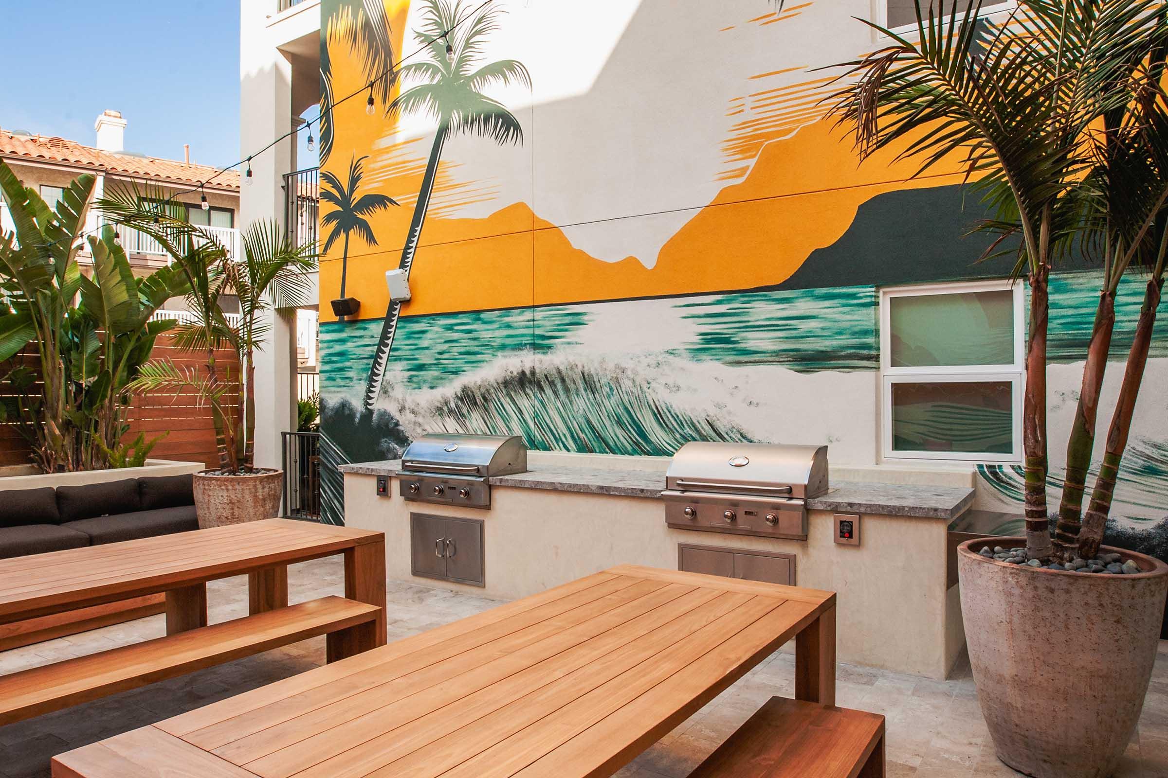 Outdoor patio with a vibrant mural featuring palm trees and ocean waves. There are two stainless steel grills and a large wooden dining table with benches. Lush greenery in decorative planters enhances the space, creating a relaxed, inviting atmosphere. Bright and colorful design elements add a lively touch.