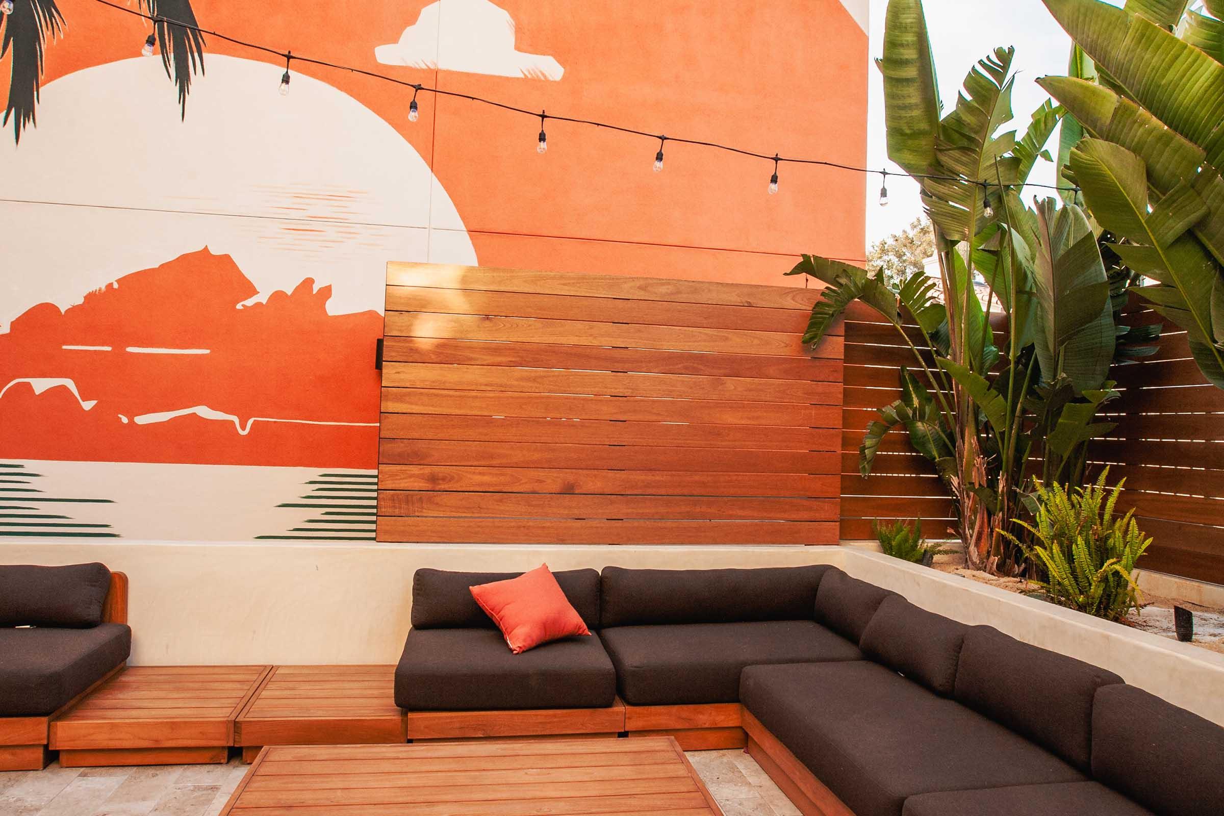 A cozy outdoor seating area featuring a dark sectional sofa with a red accent pillow, set against a vibrant orange wall with a mural of a sunset and mountains. The space is surrounded by lush greenery and wooden fencing, with string lights overhead for ambiance.