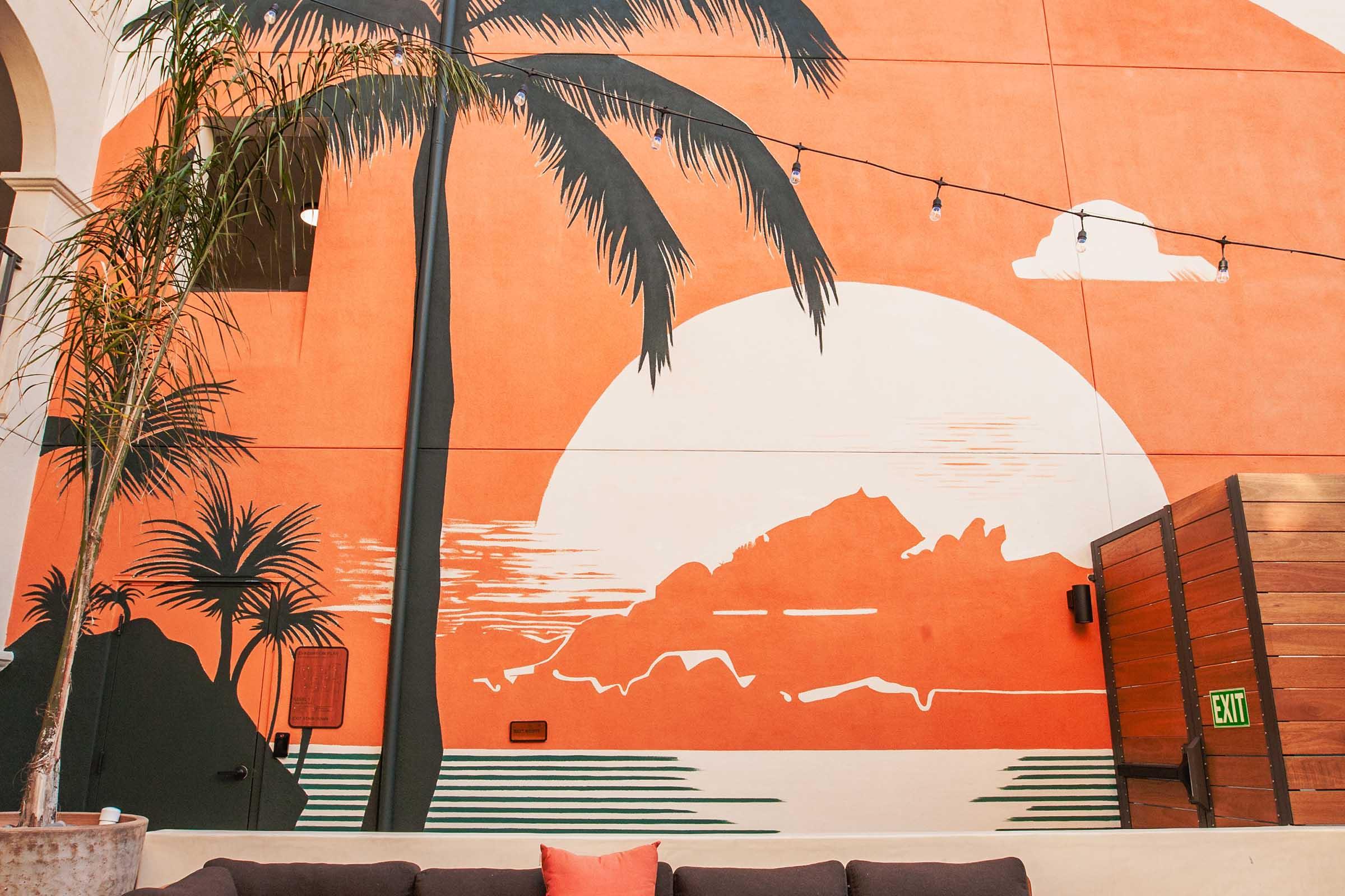 A large mural features a sunset scene with a vibrant orange background, a silhouette of mountains, and palm trees in the foreground. The design includes a stylized sun and scattered clouds, creating a tropical ambiance. A cozy seating area is visible in the foreground, enhancing the inviting atmosphere.