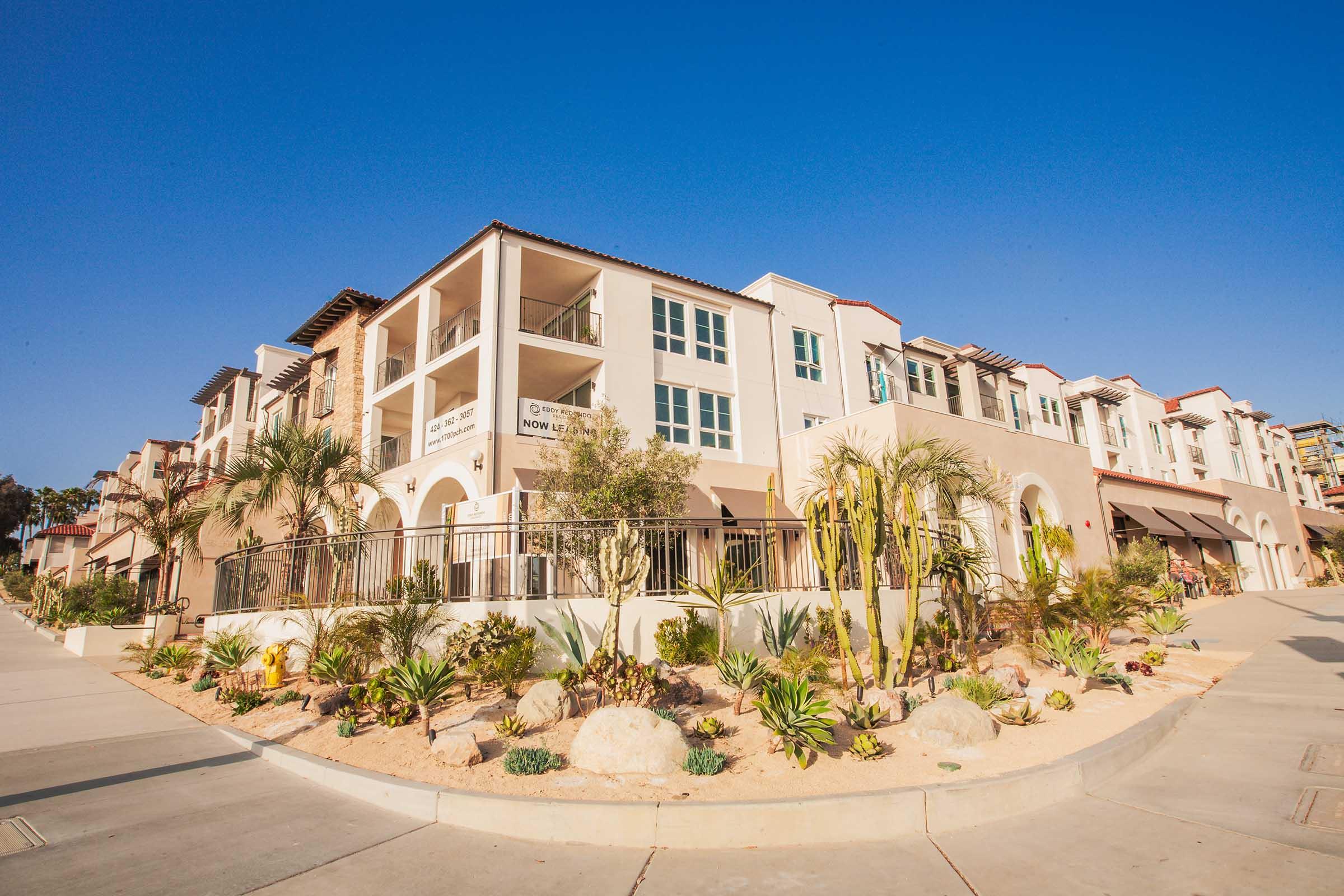 A modern residential building with a Tuscan-style facade, featuring a mix of stucco and stone elements. It has multiple balconies and large windows, surrounded by landscaped gardens with palm trees and desert plants. The scene is set under a clear blue sky, highlighting the vibrant architectural design.