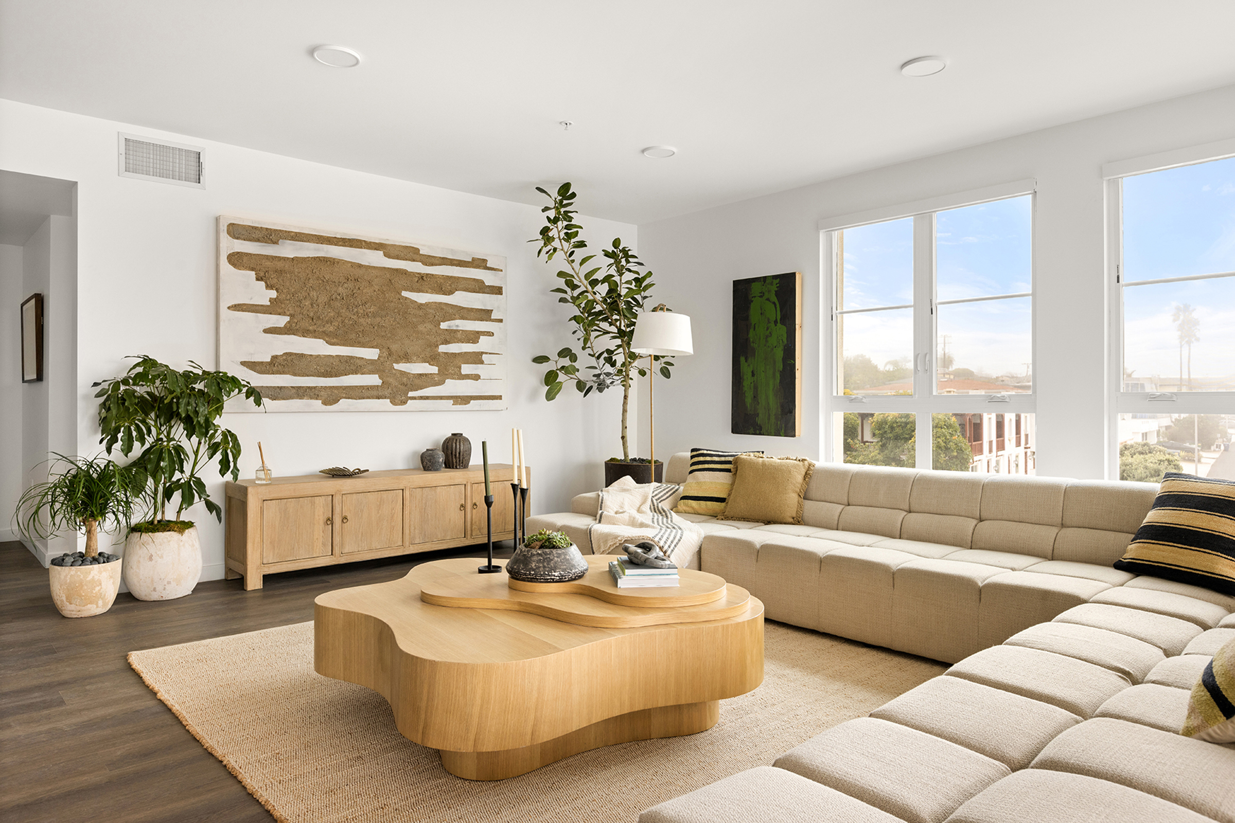 A modern living room featuring a beige sectional sofa, a unique wooden coffee table, and a light-colored rug. There are plants, decorative items on a wooden sideboard, and large windows allowing natural light. The wall is adorned with abstract art, adding a contemporary touch to the space.