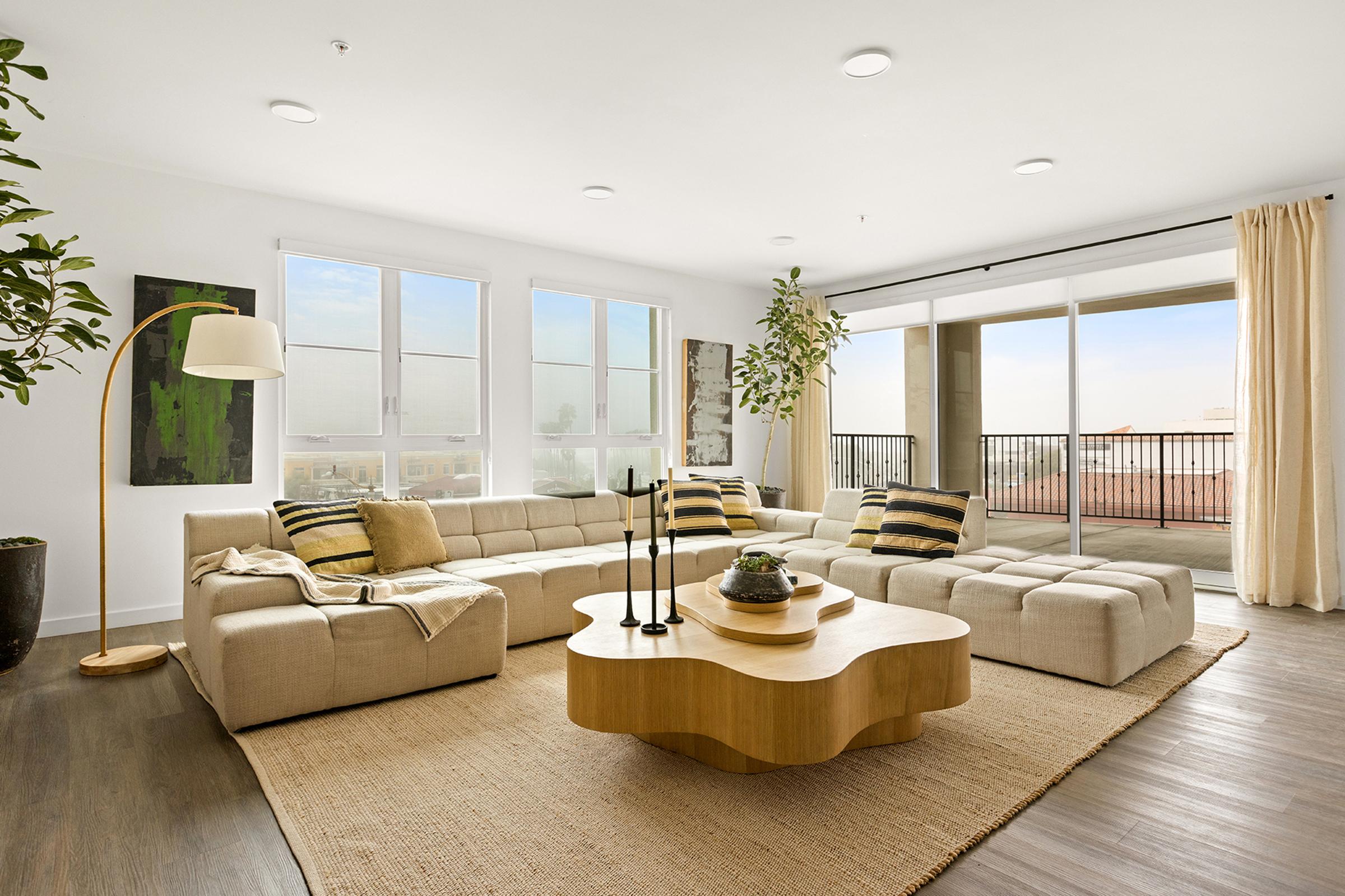 A modern living room featuring a large, beige sectional sofa with decorative pillows, a wooden coffee table, and a round area rug. There are indoor plants, a floor lamp, and large windows with sheer curtains letting in natural light, offering a view of a balcony. The decor is minimalistic and contemporary.