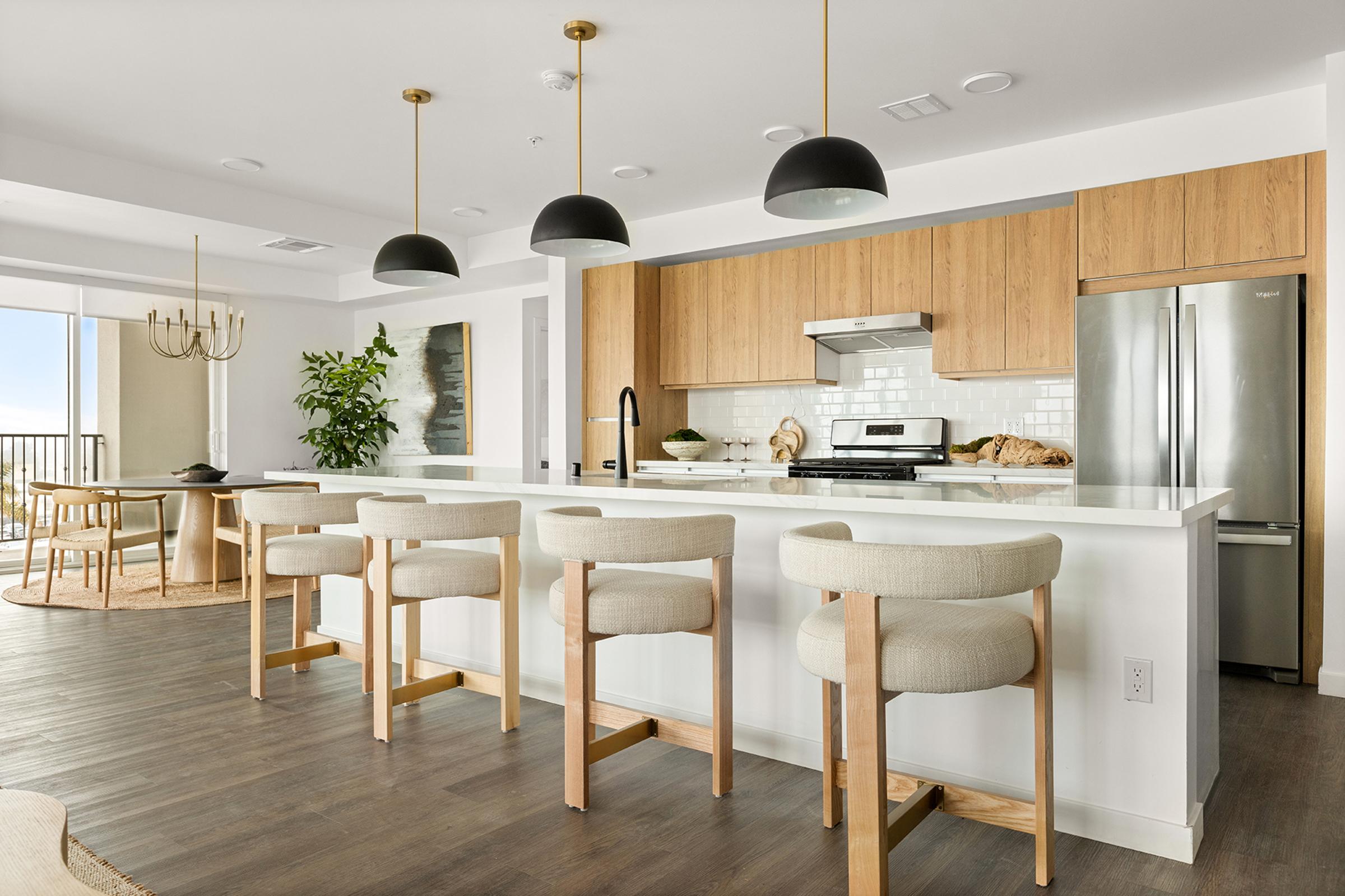 A modern kitchen featuring a large island with four light-colored stools. The cabinetry is a warm wood tone, paired with stainless steel appliances and white subway tile backsplash. Hanging pendant lights with black shades illuminate the space. A dining area with a round table and a decorative plant is visible in the background.