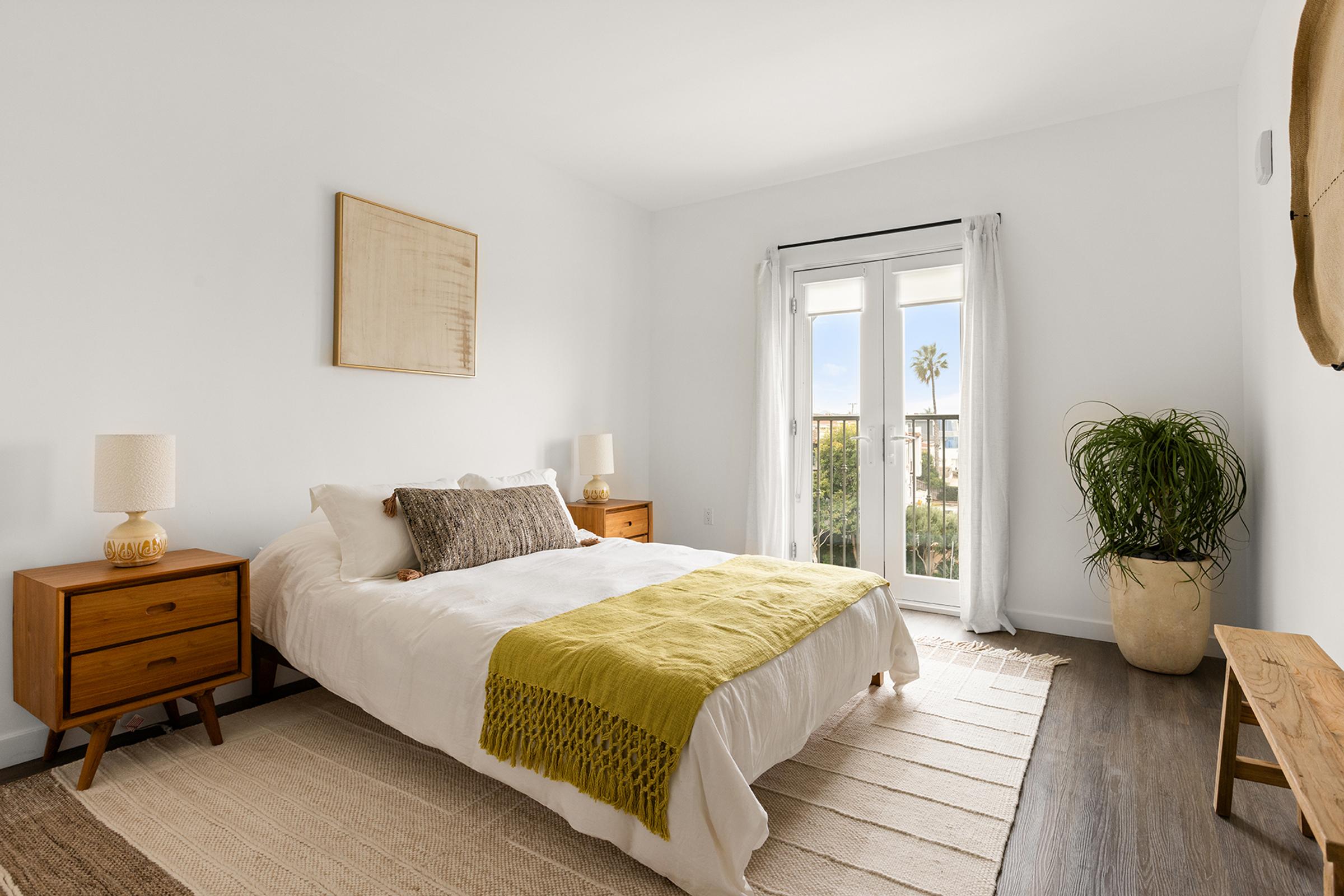 A bright and airy bedroom featuring a large bed with a white duvet and a green throw blanket, two bedside tables with lamps, and large windows that let in natural light. A potted plant adds a touch of greenery, while a textured wall hanging complements the minimalist decor.