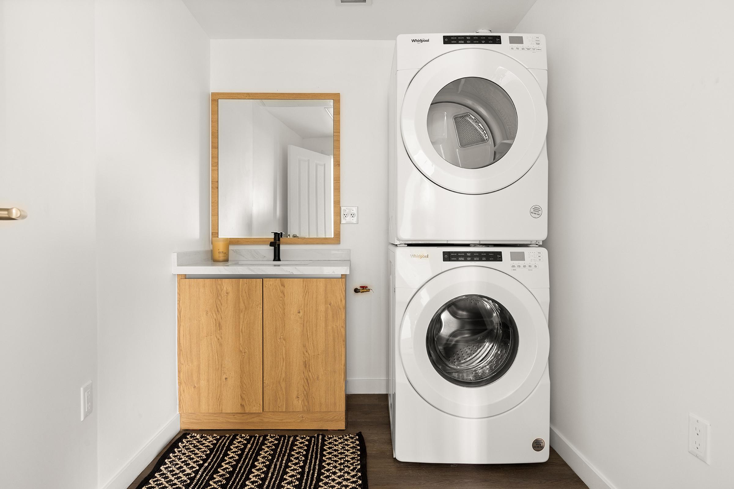 A modern laundry room featuring a stacked washer and dryer in white, a wooden cabinet with a countertop, a wall-mounted mirror, and a patterned rug on the floor. The walls are painted white, creating a bright and clean atmosphere.