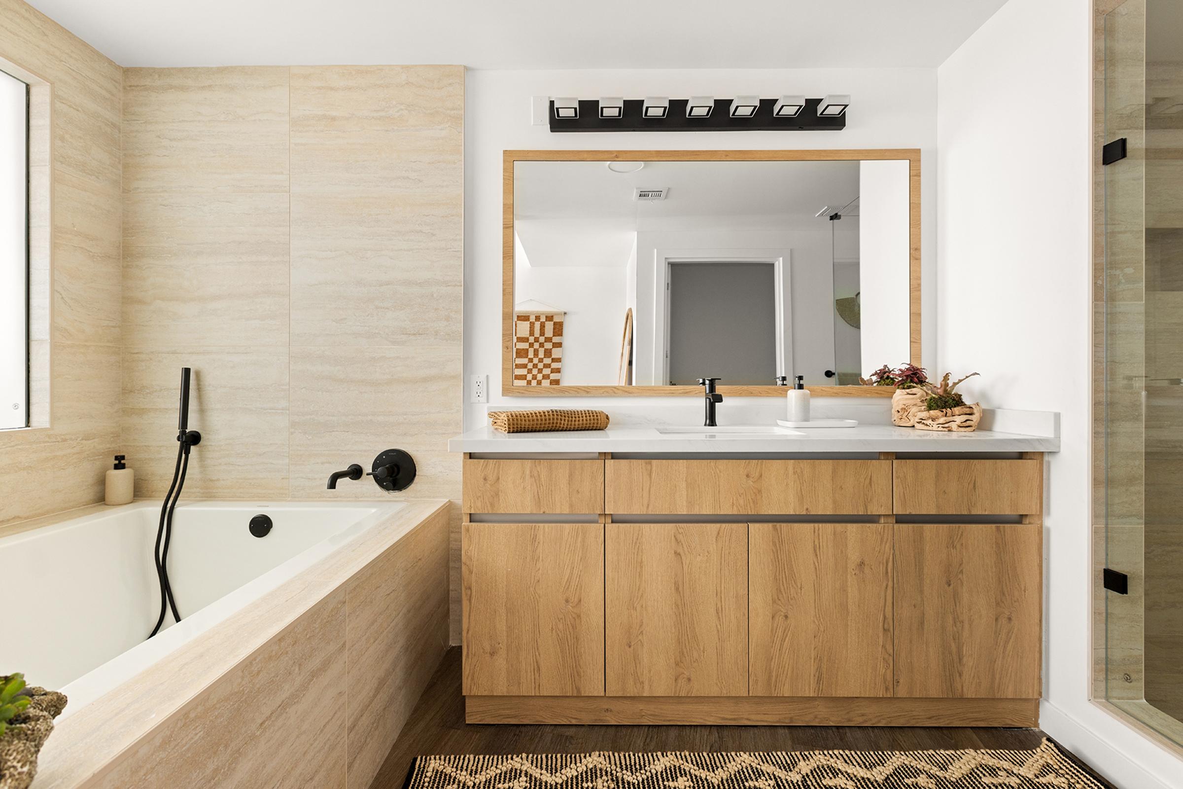 Modern bathroom featuring a freestanding bathtub, a wood vanity with black fixtures, and a large mirror. Natural elements are present with stone and wood accents. The space is well-lit and designed with a minimalist aesthetic, including a woven rug and decorative items on the counter.