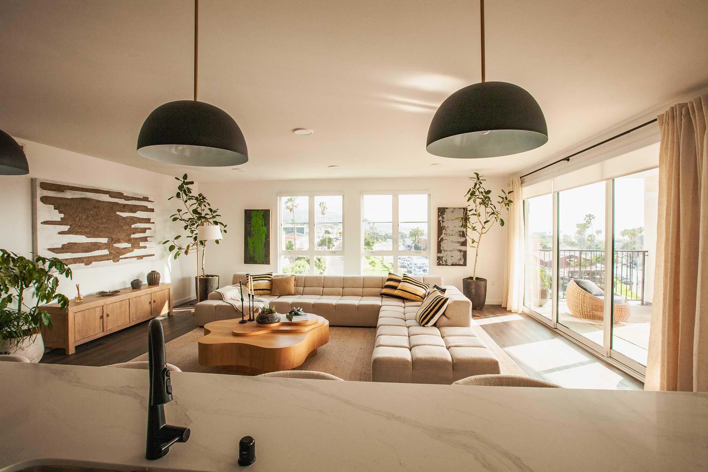 A bright and modern living room with large windows allowing natural light. The space features a light-colored sectional sofa, a wooden coffee table, and plants. A sliding glass door leads to a balcony with a view. Warm, neutral tones and stylish decor create a cozy atmosphere.