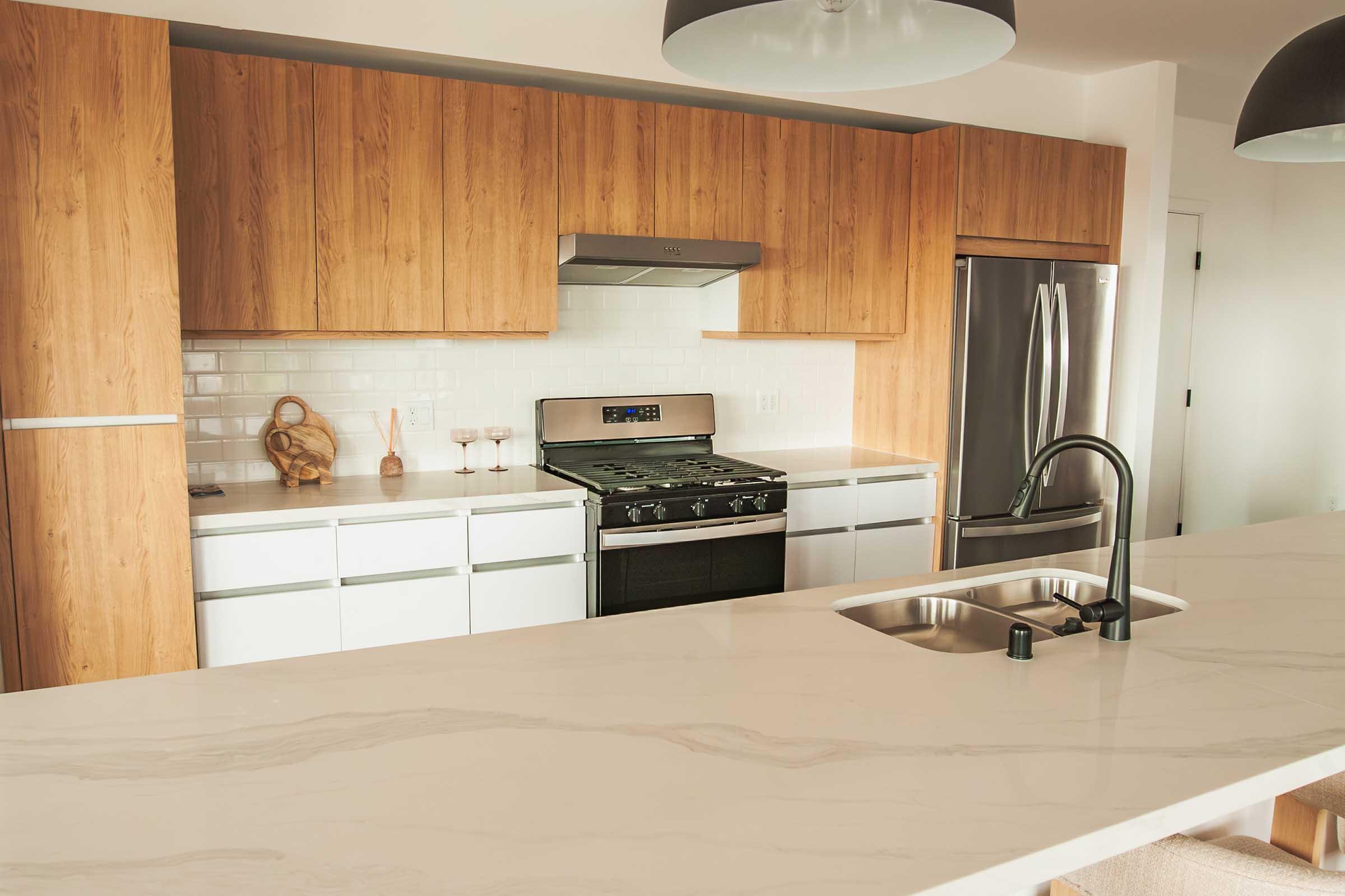 Modern kitchen featuring wooden cabinetry, stainless steel appliances, and a large marble countertop. The space includes a gas stove, a double sink with a sleek black faucet, and minimalist decor, creating a clean and inviting atmosphere.