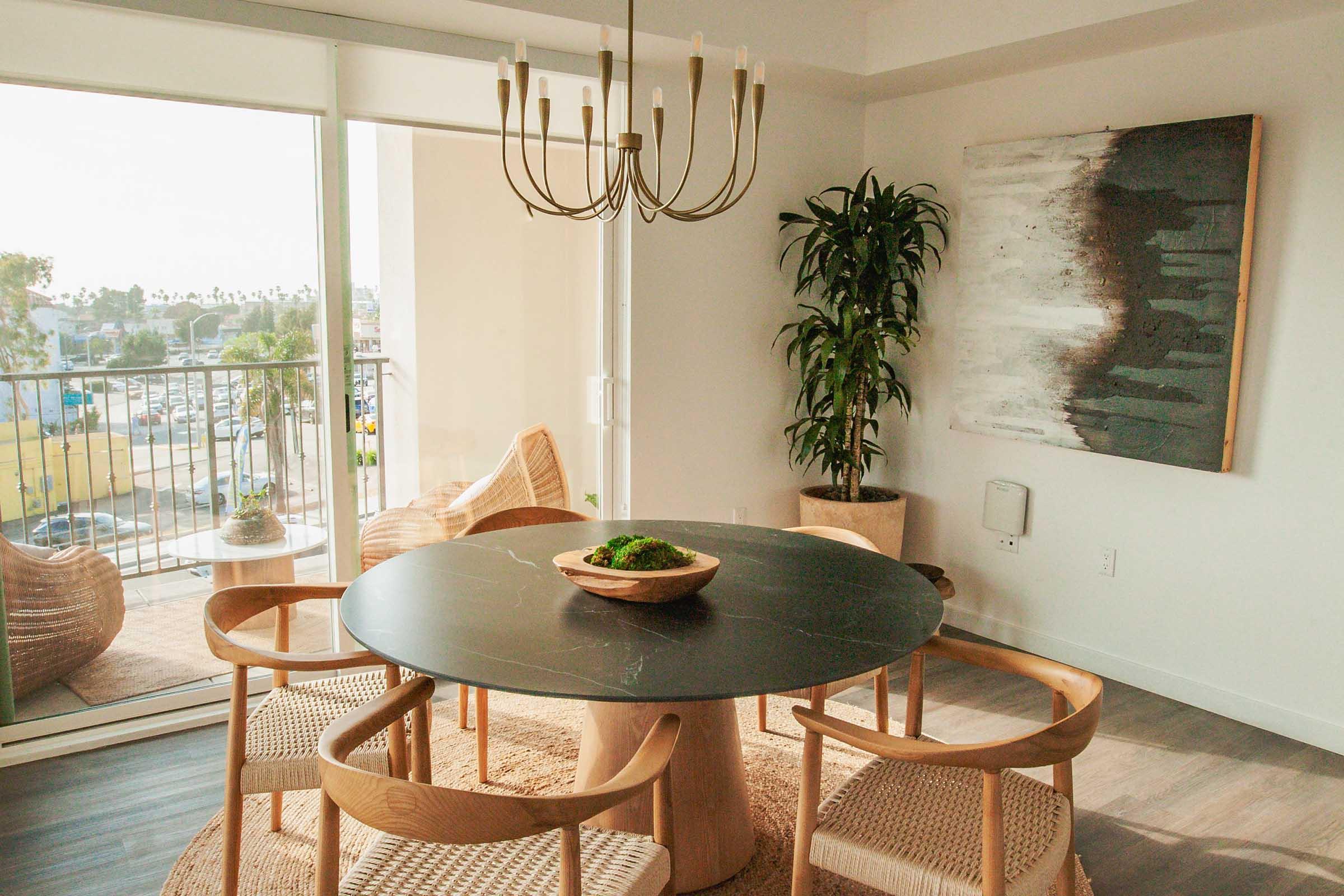 A modern dining area featuring a round black table with wooden chairs and a bowl of greenery on top. The room has large windows that allow natural light, showcasing a view outside. A stylish chandelier hangs above the table, and there's a potted plant in the corner. The walls are decorated with contemporary artwork.