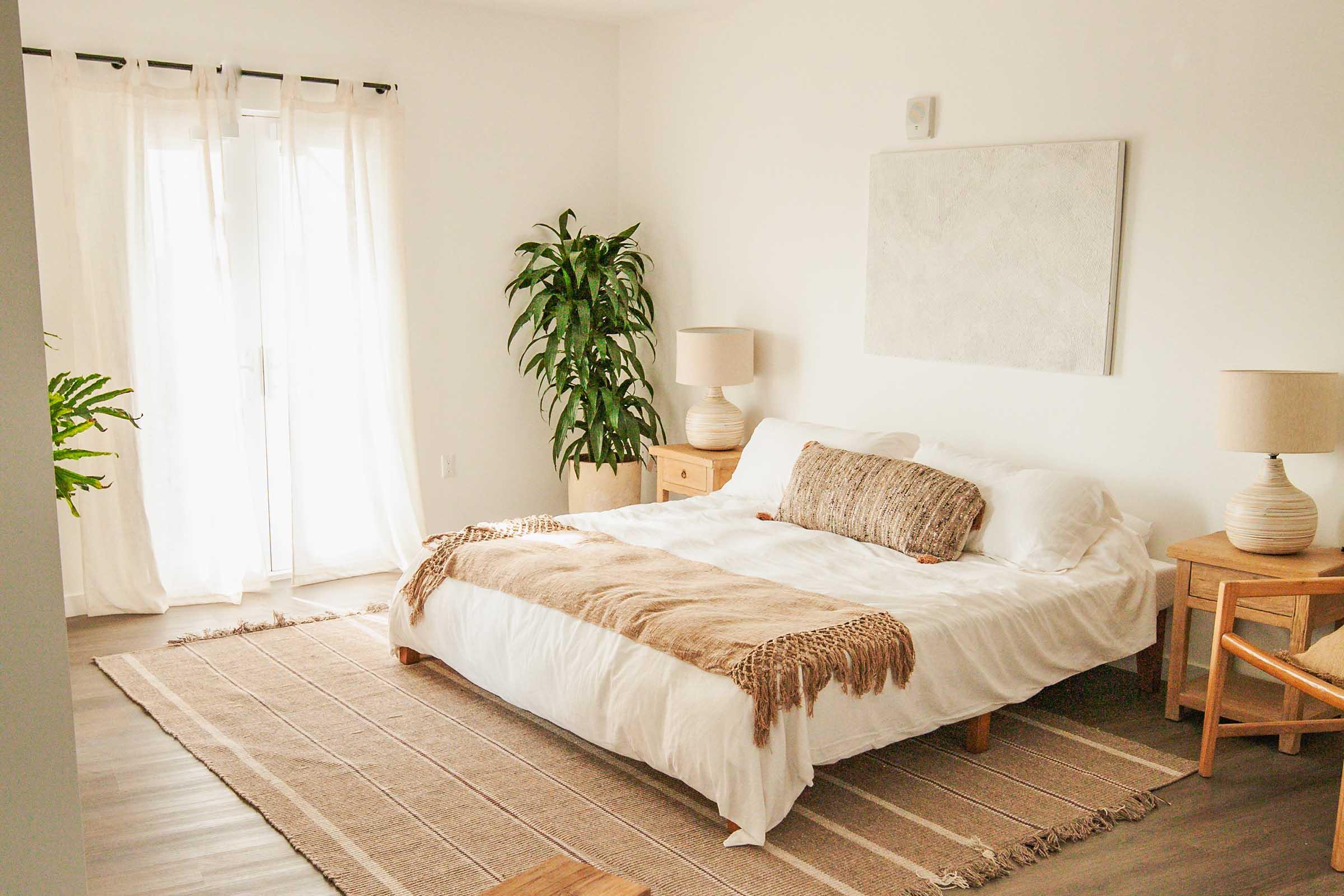 A bright, minimalistic bedroom featuring a large bed with white bedding, a decorative throw, and wooden nightstands. Two lamps sit on the nightstands, and a large window with sheer curtains lets in natural light. A green plant stands in the corner, adding a touch of greenery to the serene space.