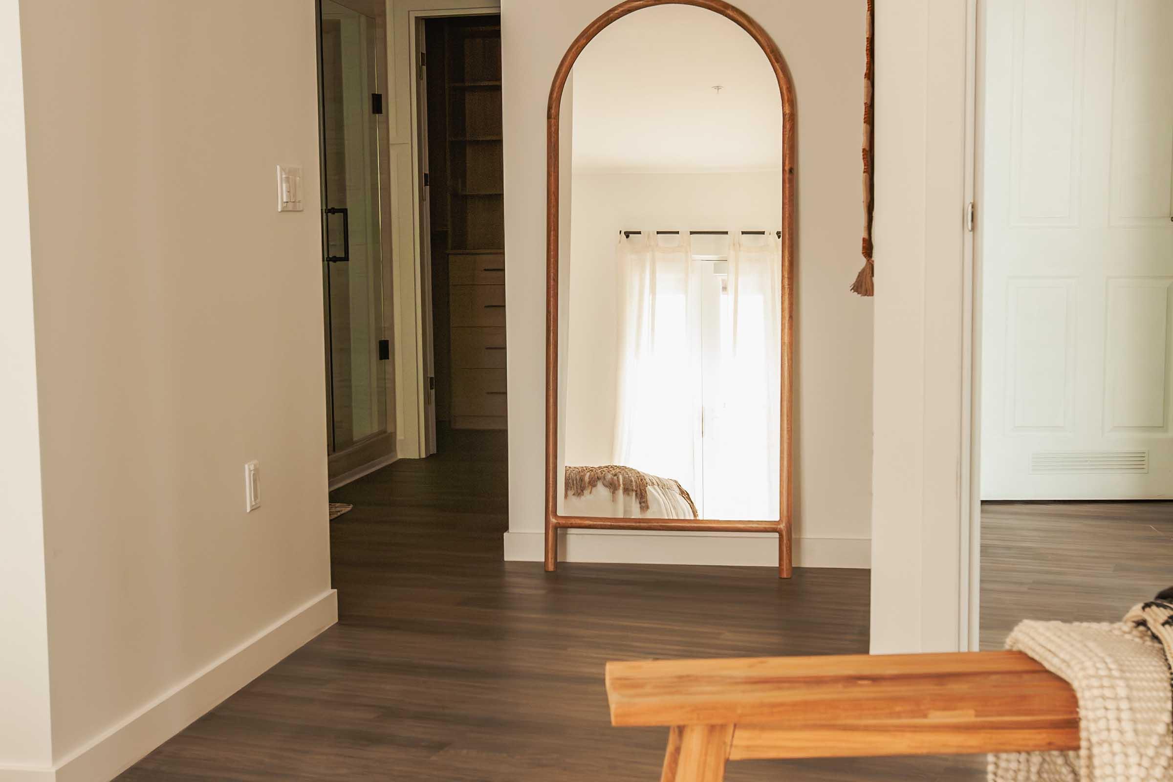 A spacious, modern interior featuring a large, arched mirror framed in wood. The room has warm, neutral-colored walls and wooden flooring. Natural light filters through sheer curtains, illuminating the space. A glimpse of a bathroom door and closet can be seen in the background, and a wooden chair is partially visible in the foreground.