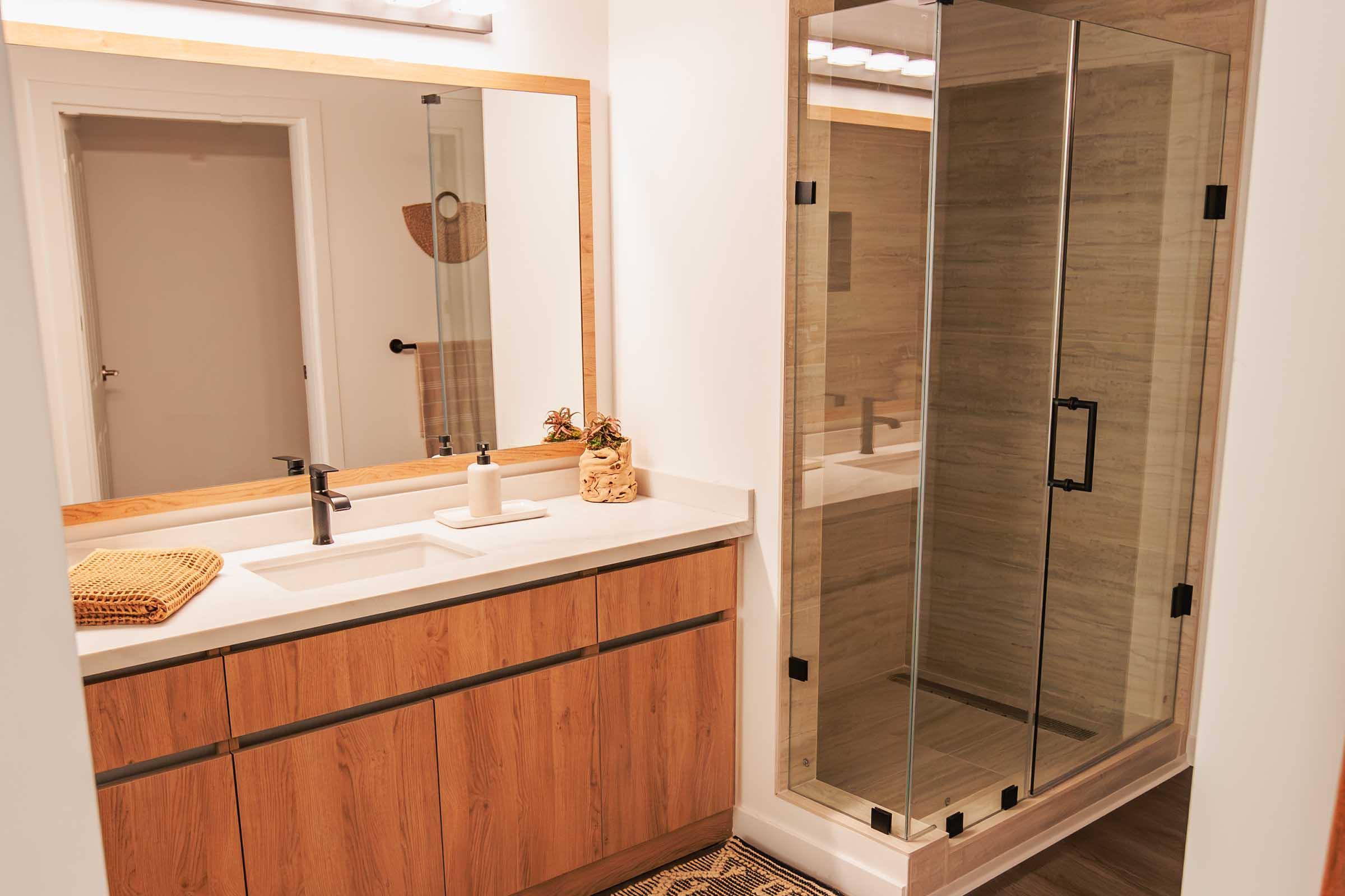 Modern bathroom featuring a wooden vanity with a double sink and stylish faucet. A large mirror hangs above the sink. To the right, a glass shower enclosure with minimalistic design. Soft lighting highlights the neutral color palette, creating a clean and inviting atmosphere. A woven mat complements the decor.