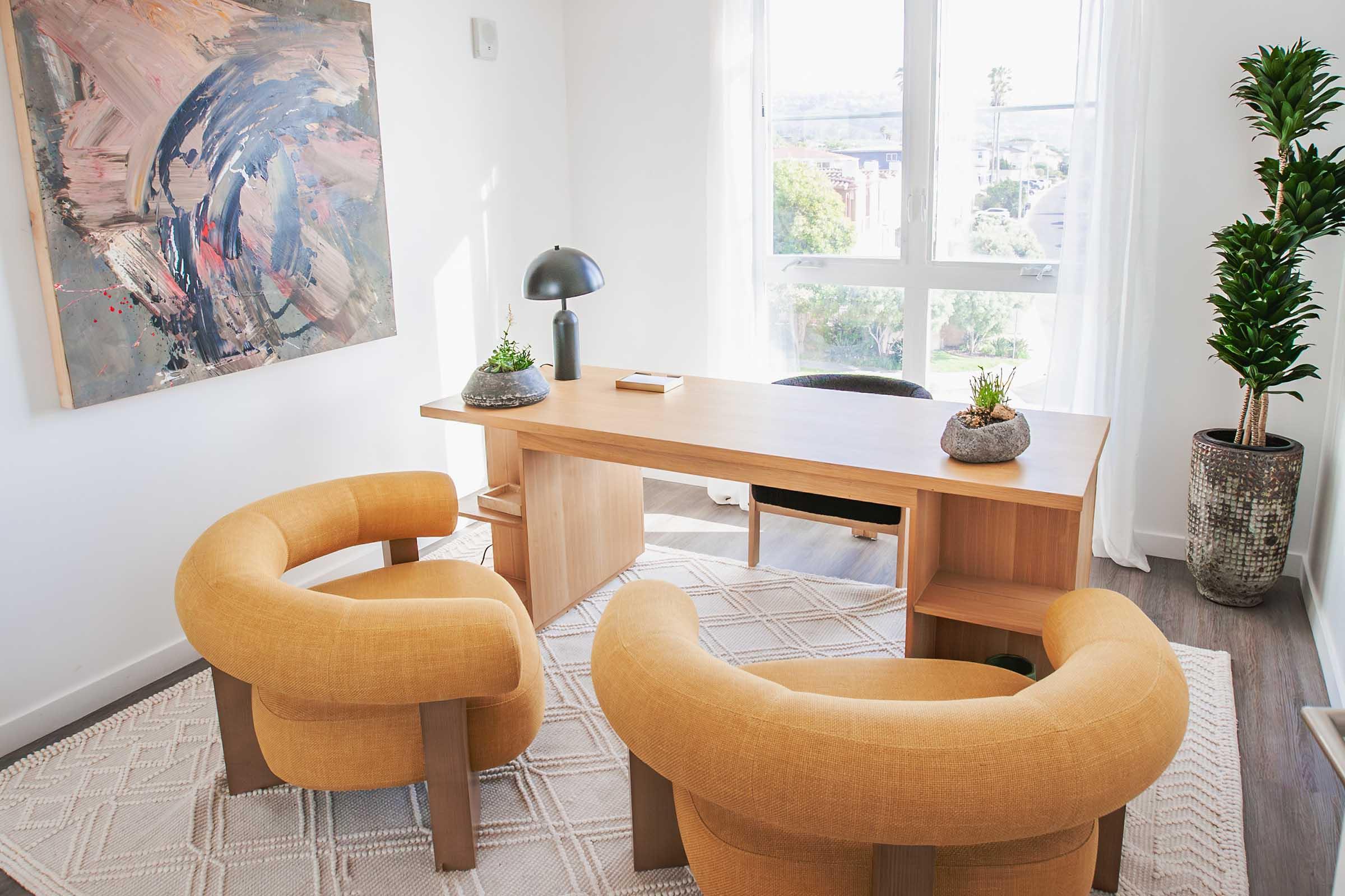 A modern office space featuring a wooden desk, two round yellow chairs, and a decorative rug. There is a lamp on the desk, small plants on both the desk and in pots beside a window, and a colorful abstract painting on the wall, adding a stylish touch to the serene environment.
