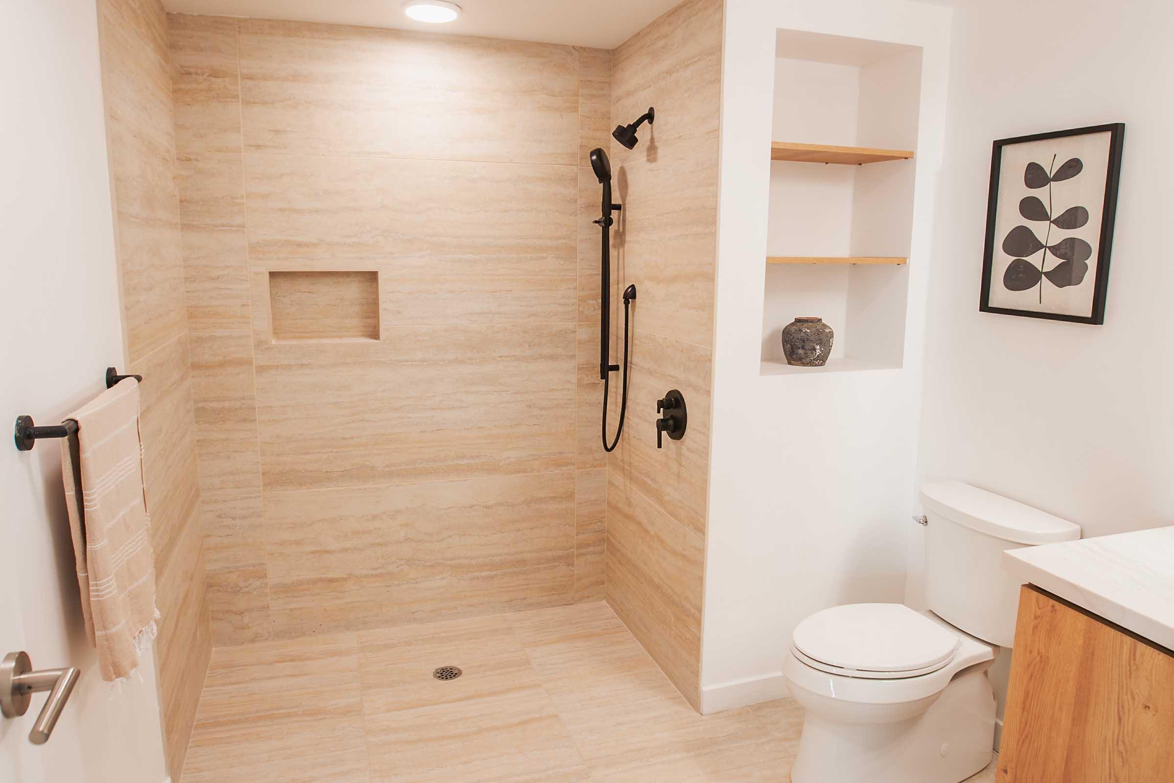 A modern bathroom featuring a spacious walk-in shower with tiled walls, a rainfall showerhead, and a built-in shelf. There is a white toilet and a wooden vanity. Shelves on the wall above the shower hold decorative items, and a framed art piece is mounted beside the shelves. The overall design is clean and minimalist.