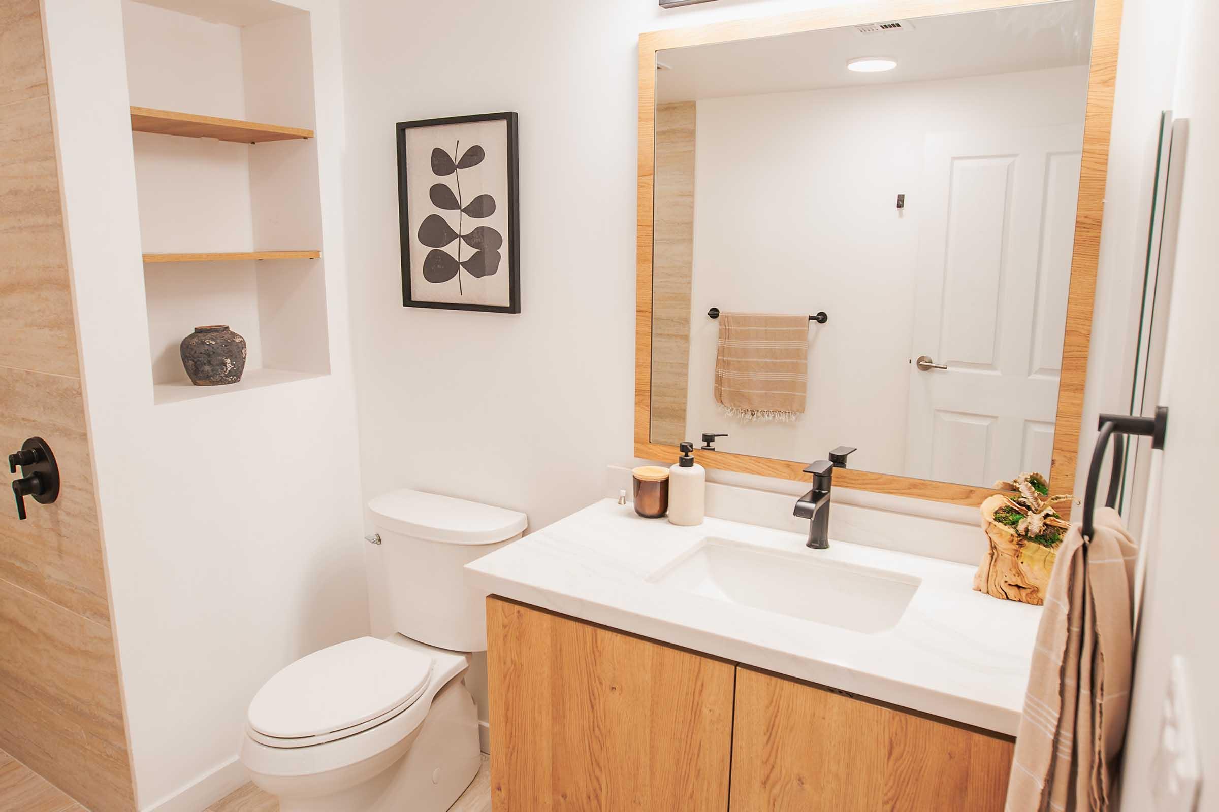 A modern bathroom featuring a white toilet, wooden vanity with a marble countertop, and a wall-mounted mirror. There is a framed leaf print on the wall, a decorative vase on a shelf, and a towel hanging nearby. The decor combines natural wood tones with a clean, minimalist design.