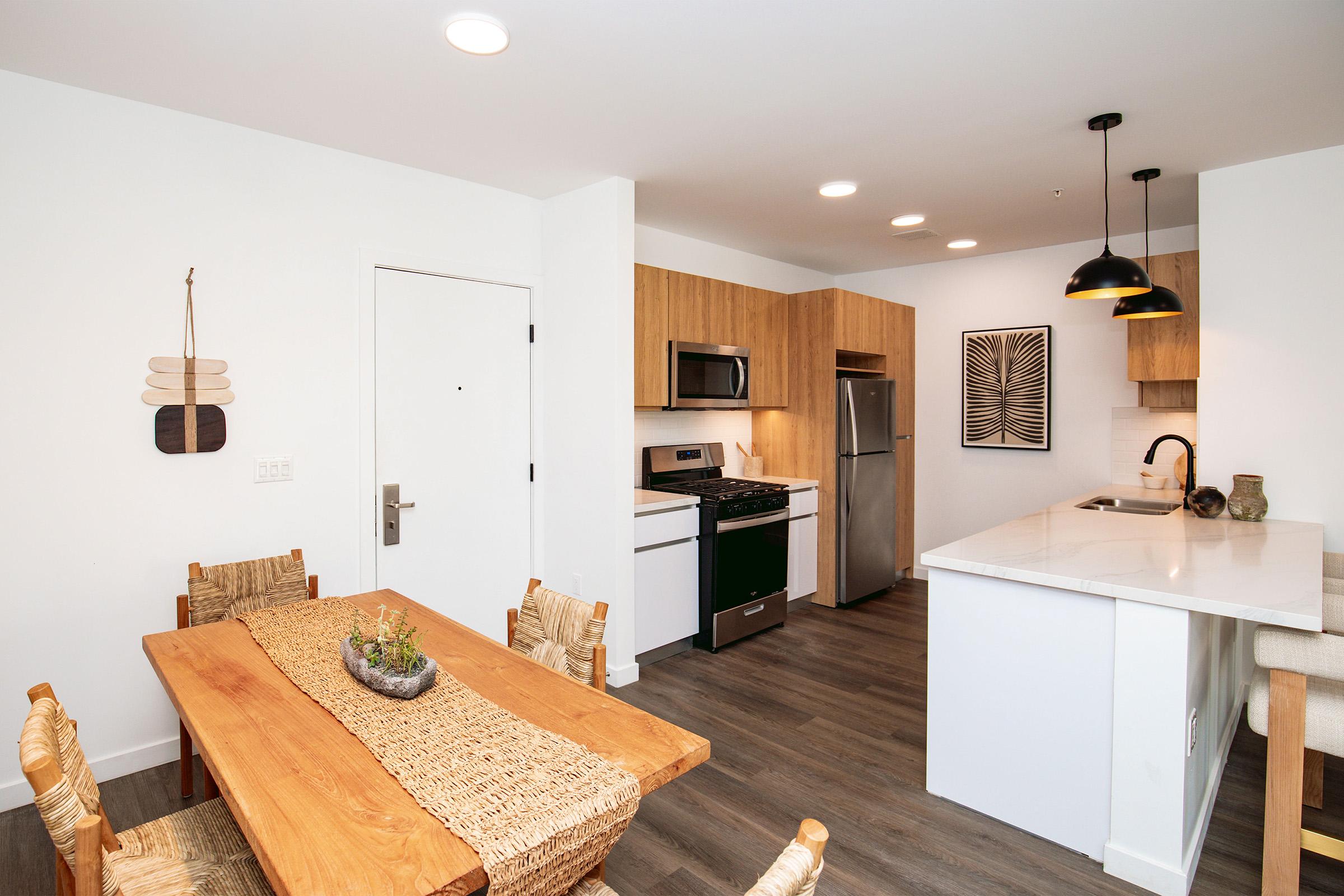 Modern kitchen and dining area featuring a wooden table with woven seating, a sleek kitchen island with bar seating, stainless steel appliances, and light wood cabinetry. White walls and contemporary pendant lights create a bright, inviting atmosphere.