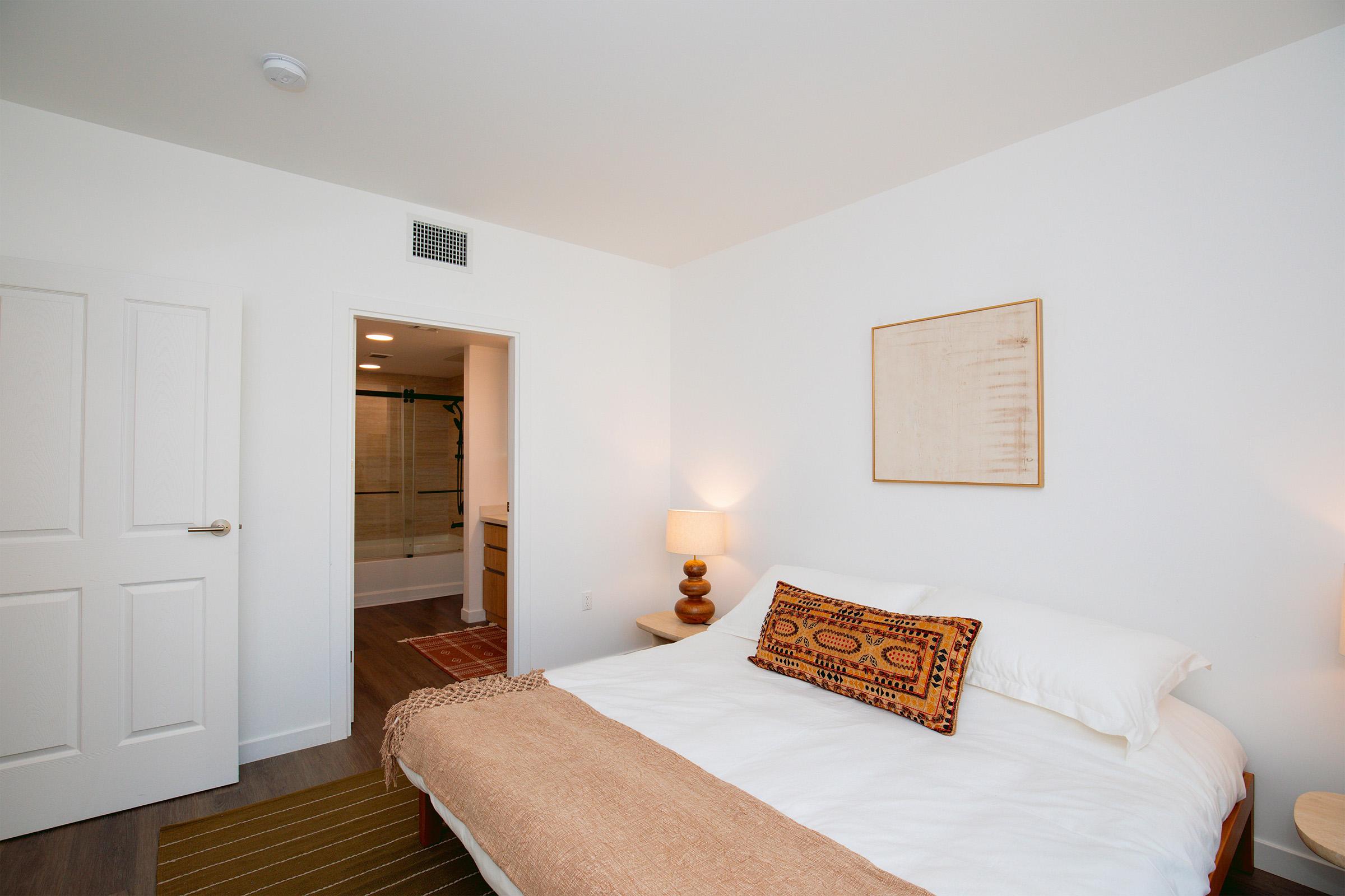 A neatly arranged bedroom featuring a white bed with decorative pillows and a warm-colored throw. There are two bedside lamps on wooden tables, a textured wall art piece, and a door leading to a bathroom. The flooring is light wood, and a patterned area rug adds warmth to the room.