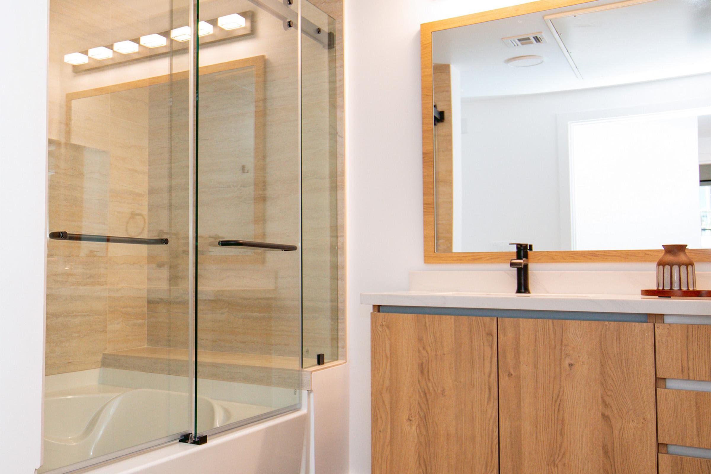 A modern bathroom featuring a glass shower enclosure and a wooden vanity with a sink. The decor includes a large mirror with warm lighting above. Light-colored tiles line the shower area, and the overall design is sleek and minimalist.