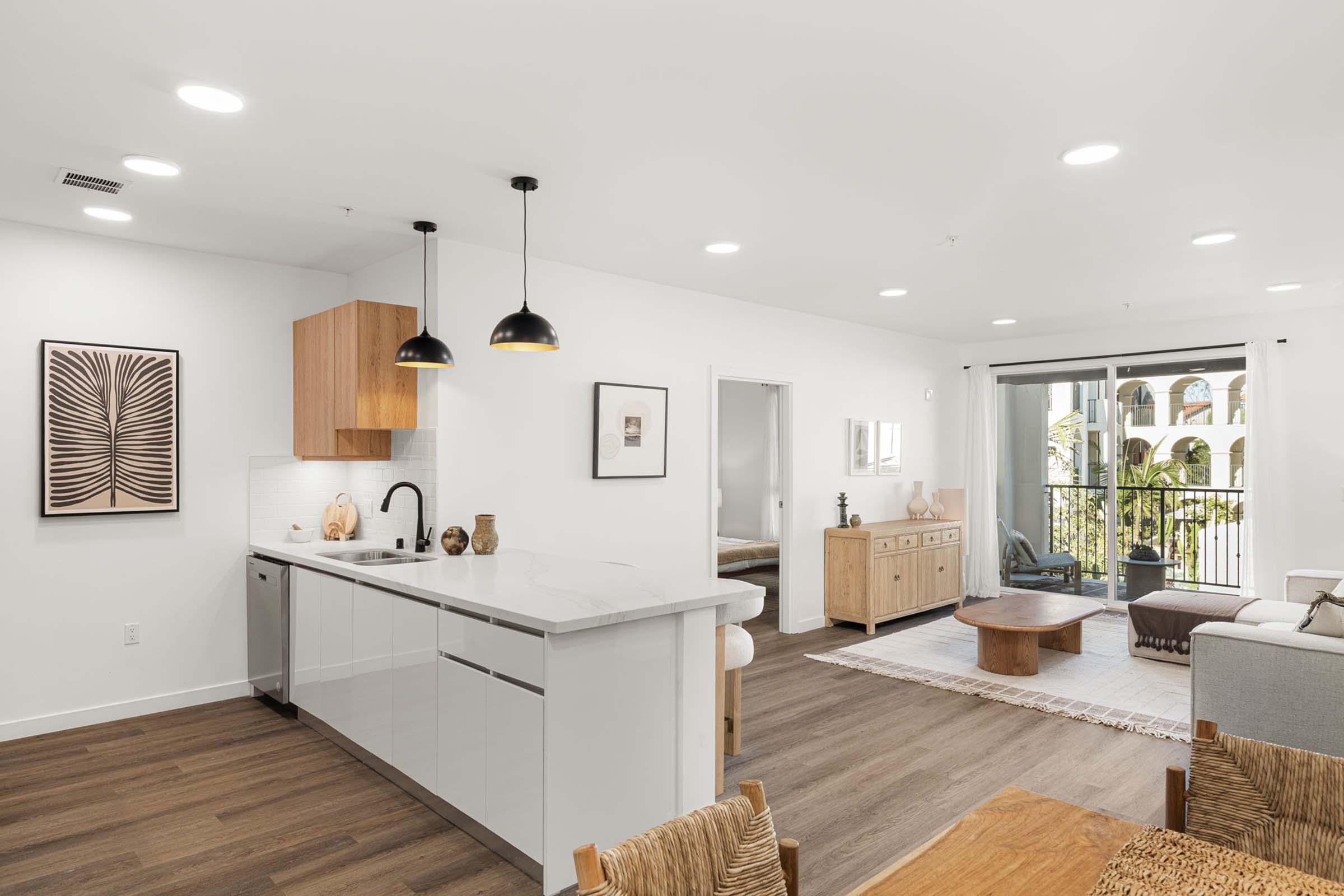 Modern open-concept kitchen and living area featuring sleek cabinetry, a stylish island with pendant lighting, and a dining table. Natural light floods the space through large windows, showcasing a balcony view. Minimalist decor enhances the contemporary aesthetic.