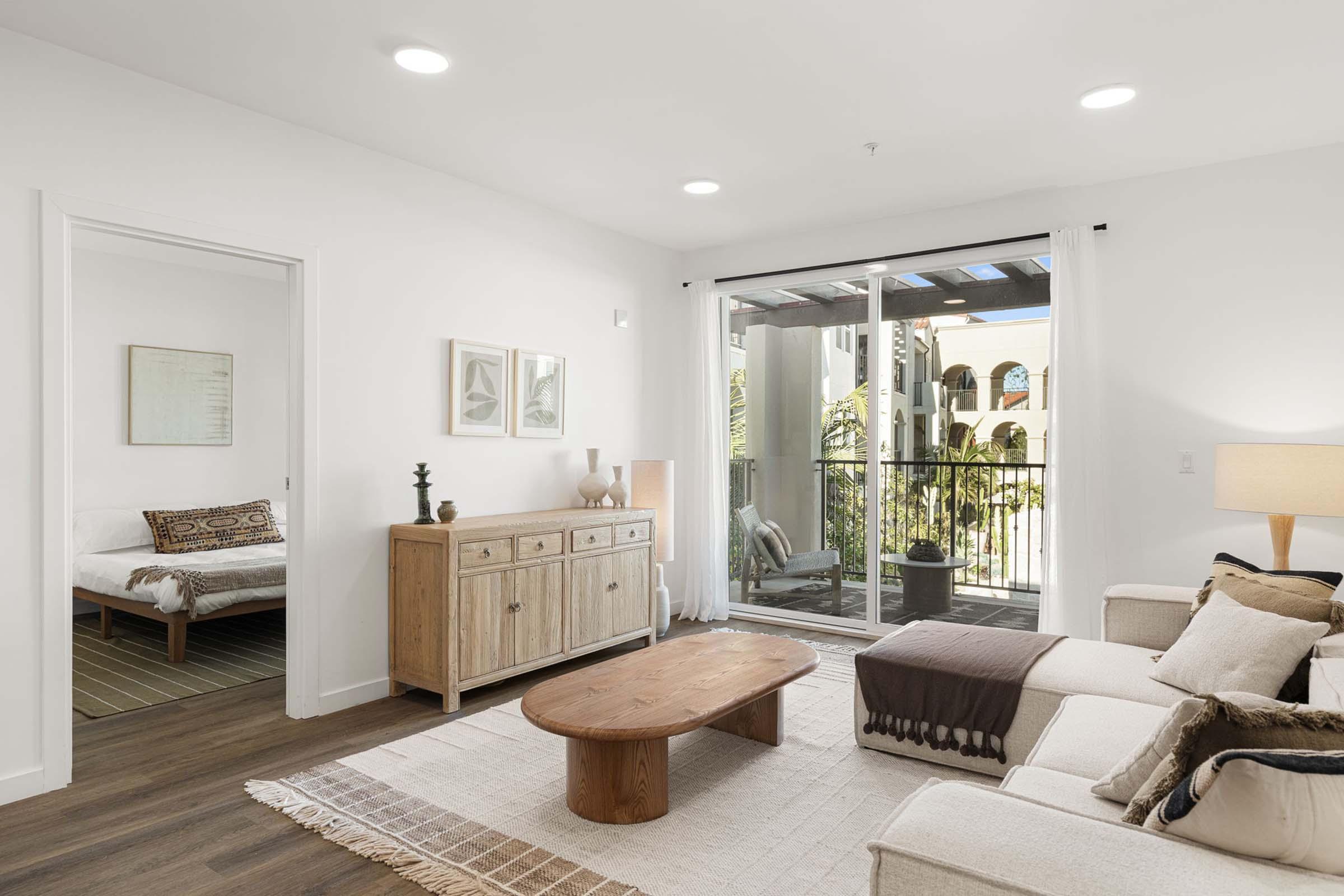 A modern living room with a light-colored sofa, wooden coffee table, and sideboard. Natural light streams in through a glass door leading to a balcony, which features plants. A cozy bedroom with a patterned bedspread is visible through an open door, complemented by neutral decor and warm lighting.
