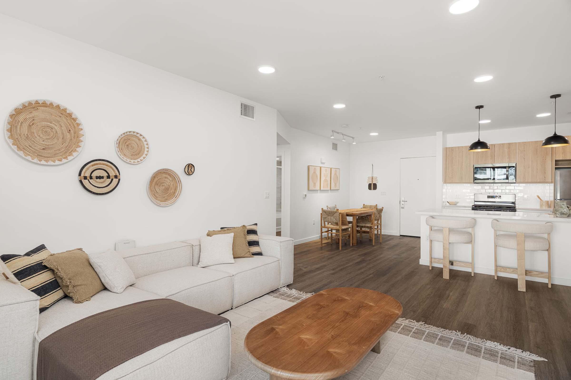 A modern living room featuring a light-colored sofa with decorative cushions, a wooden coffee table, and wall art made from circular woven materials. In the background, there's a dining area with a wooden table and chairs, and a kitchen with a sleek design, including cabinets and appliances.