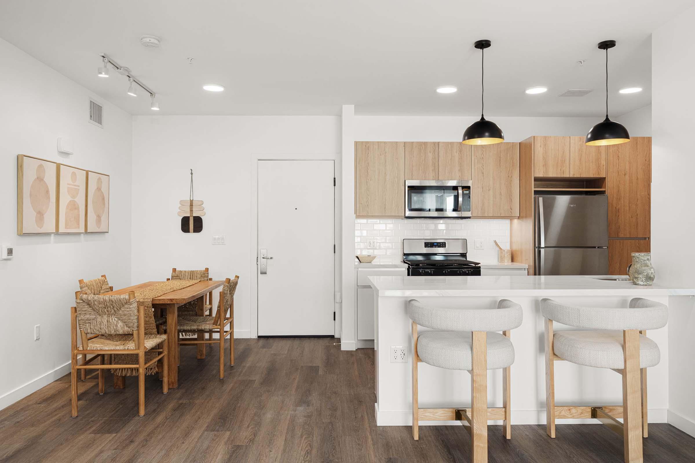 A modern kitchen and dining area featuring a wooden dining table with chairs, an open layout with a white counter and bar stools, and a kitchen equipped with stainless steel appliances. The walls are decorated with framed artwork, and there is natural light illuminating the space.