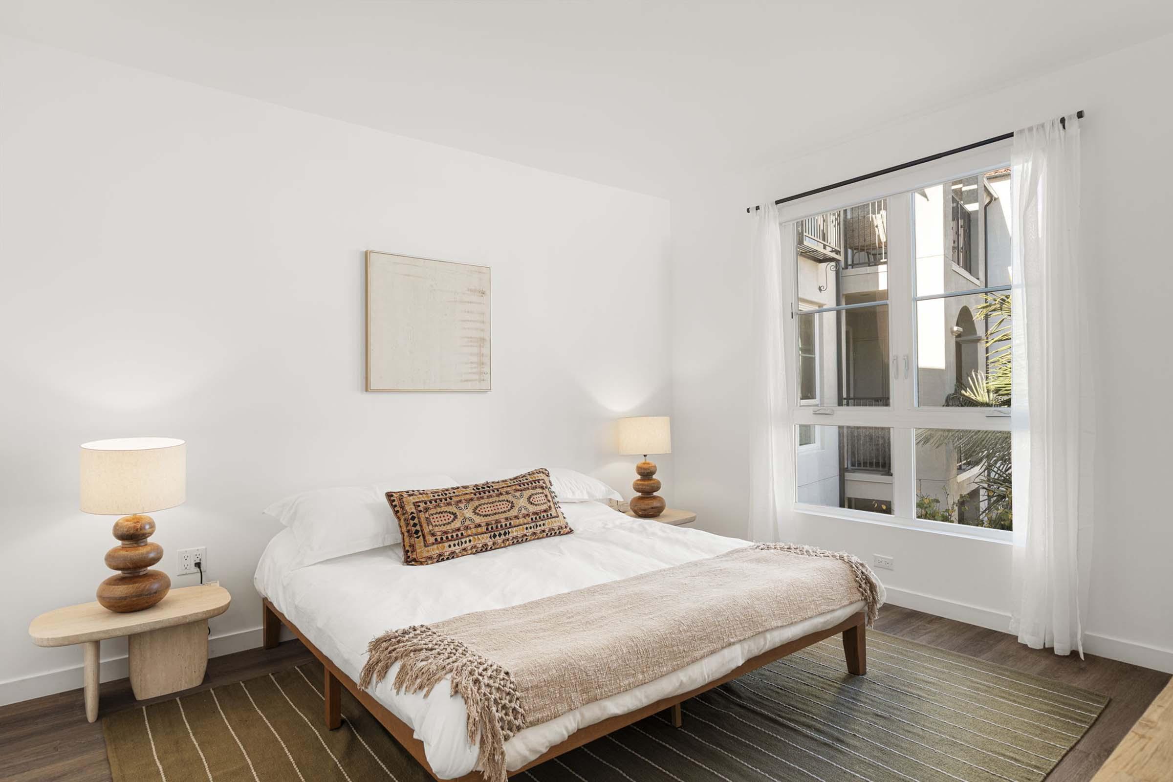 A bright, modern bedroom featuring a large bed with a white duvet and decorative throw, two bedside lamps with wooden bases, and a small wooden nightstand. There's a window with sheer curtains allowing natural light, and a simple abstract artwork on the wall. The floor has a textured rug.