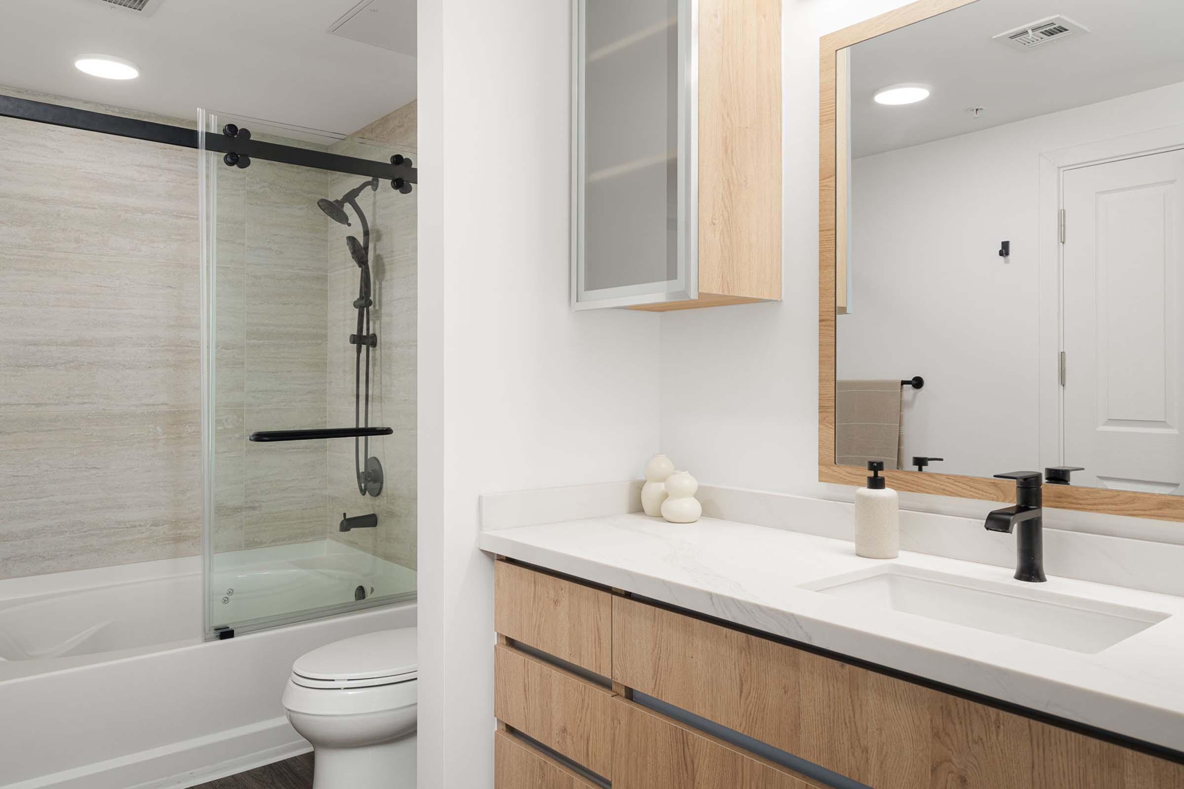 Modern bathroom featuring a glass shower enclosure with a black frame, a white bathtub, a stylish wooden vanity with a white countertop, a black faucet, and minimalistic decor items. The walls have a light beige tone, and a large mirror reflects the space.