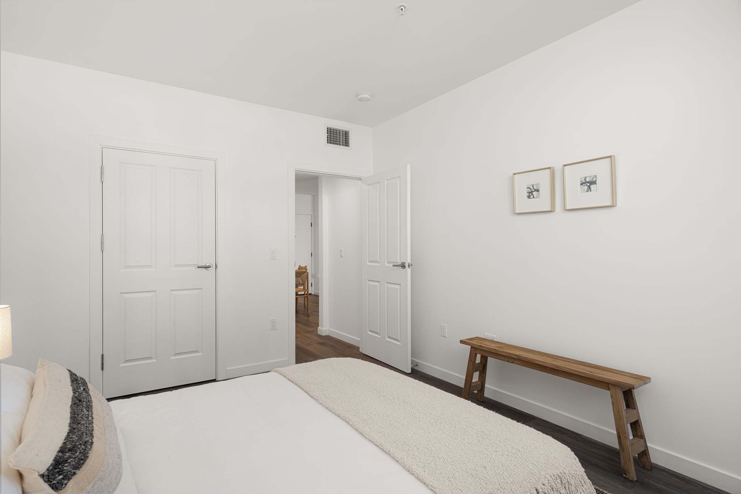 A minimalist bedroom featuring a bed with light bedding, a simple wooden bench at the foot, and a white wall with two framed pictures. A door leads to another room, and there is a small window above the bed. The flooring is dark wood, creating a modern and clean aesthetic.