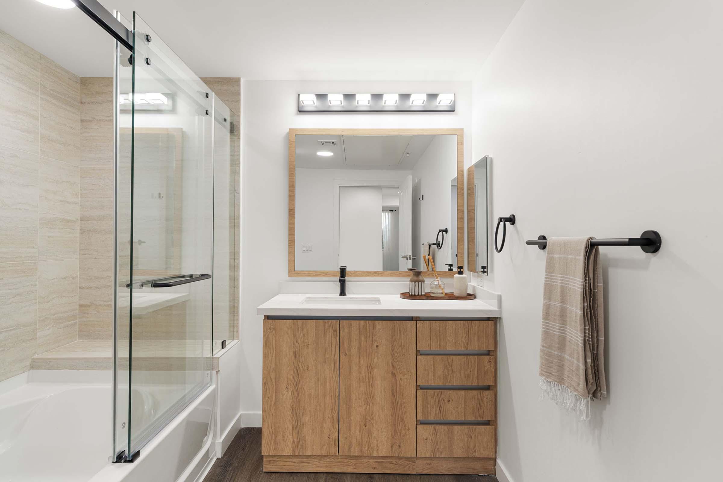A modern bathroom featuring a glass shower enclosure, a bathtub, and a wooden vanity with a white sink. The vanity is complemented by a large mirror and stylish light fixtures. A towel hangs on a black fixture, and there is a neutral-colored towel on the vanity, creating a clean, contemporary look.