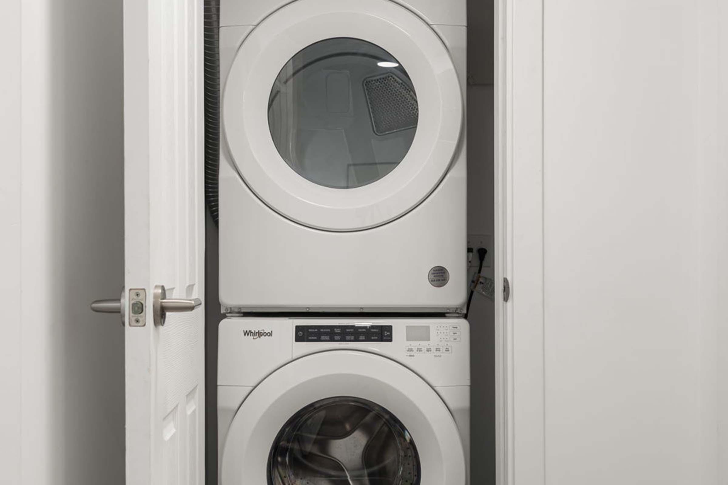 A stacked washer and dryer unit in a closet. The washer is at the bottom with a control panel displayed, while the dryer is on top with a large circular door. The interior of the closet is white, with a door slightly ajar.