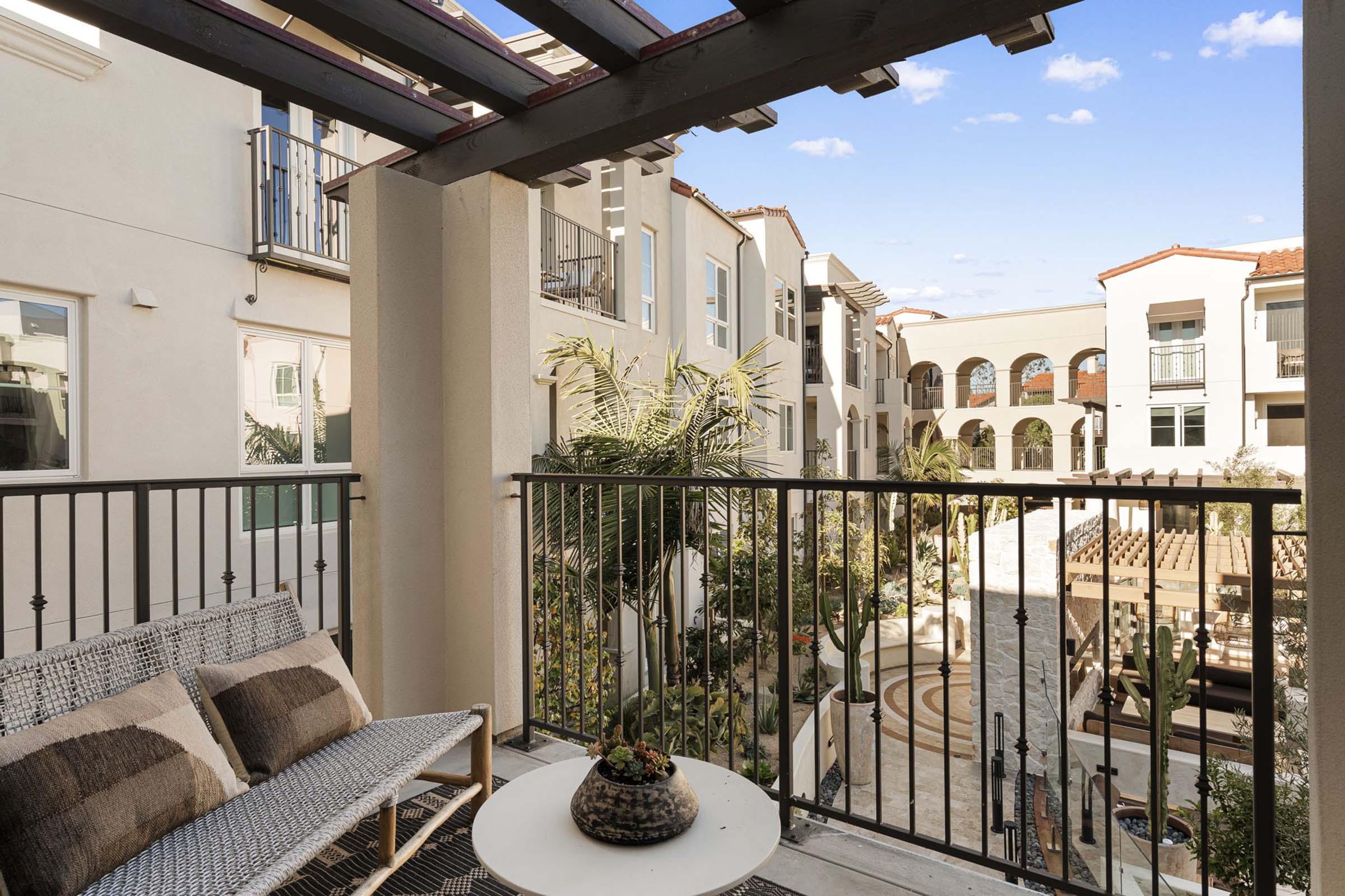 View from a balcony overlooking a courtyard with modern architecture. The scene features a seating area with a small table and potted plant, surrounded by lush greenery and palm trees. The background showcases multi-level buildings with arches and a clear blue sky.