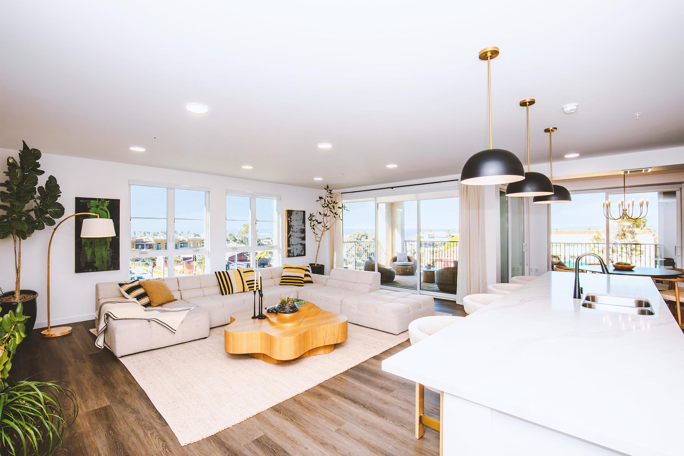 A modern living room featuring a light-colored sectional sofa, a wooden coffee table, and a large window with scenic views. The space includes indoor plants, stylish pendant lights, and a dining area with elegant decor. The walls are bright, enhancing the airy atmosphere.
