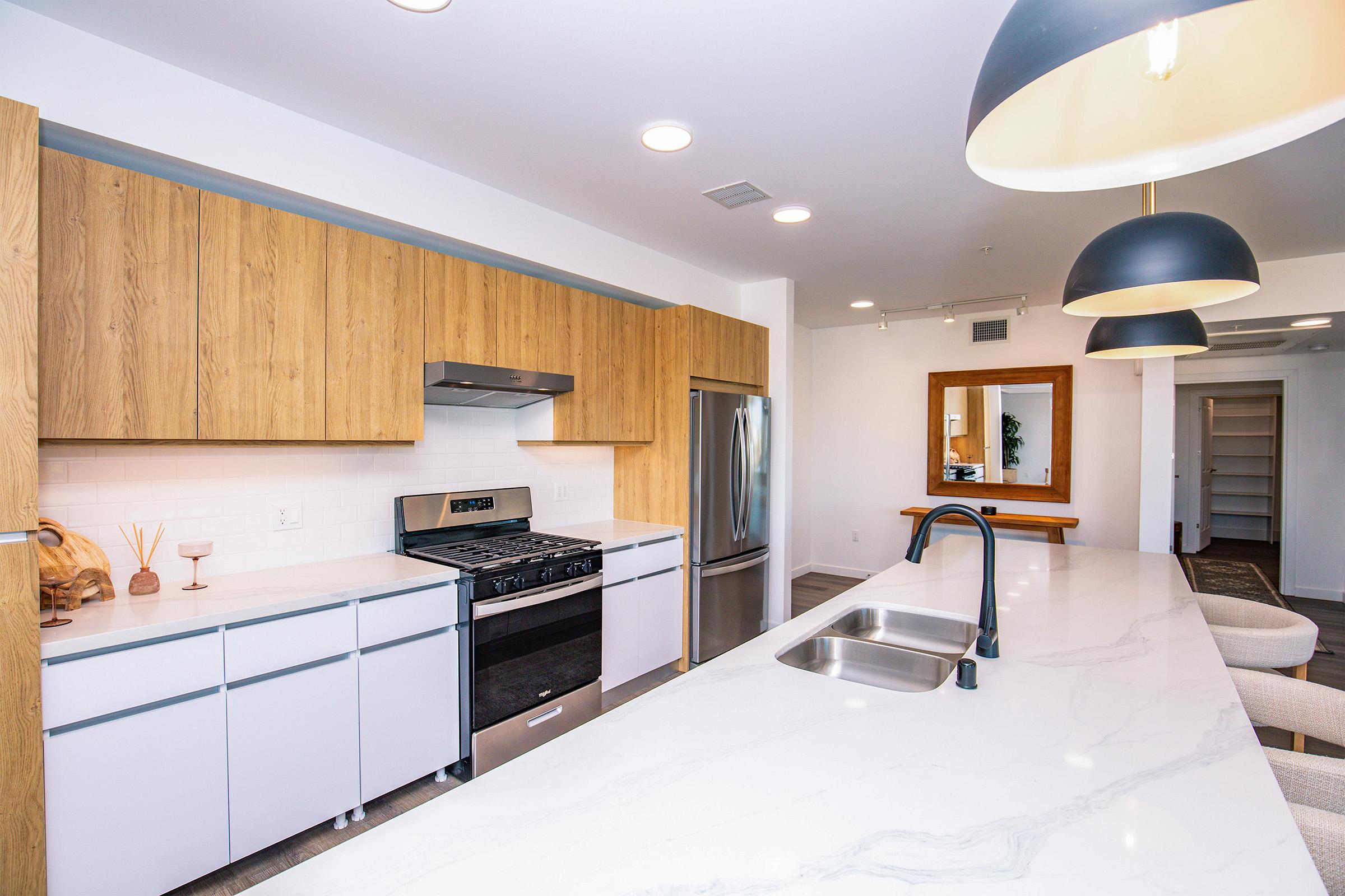 Modern kitchen featuring a large island with a sink, sleek white cabinetry, wooden upper cabinets, stainless steel appliances, and bright overhead lights. The space is well-lit with natural light and includes a large mirror in the background. The overall design is contemporary and inviting.