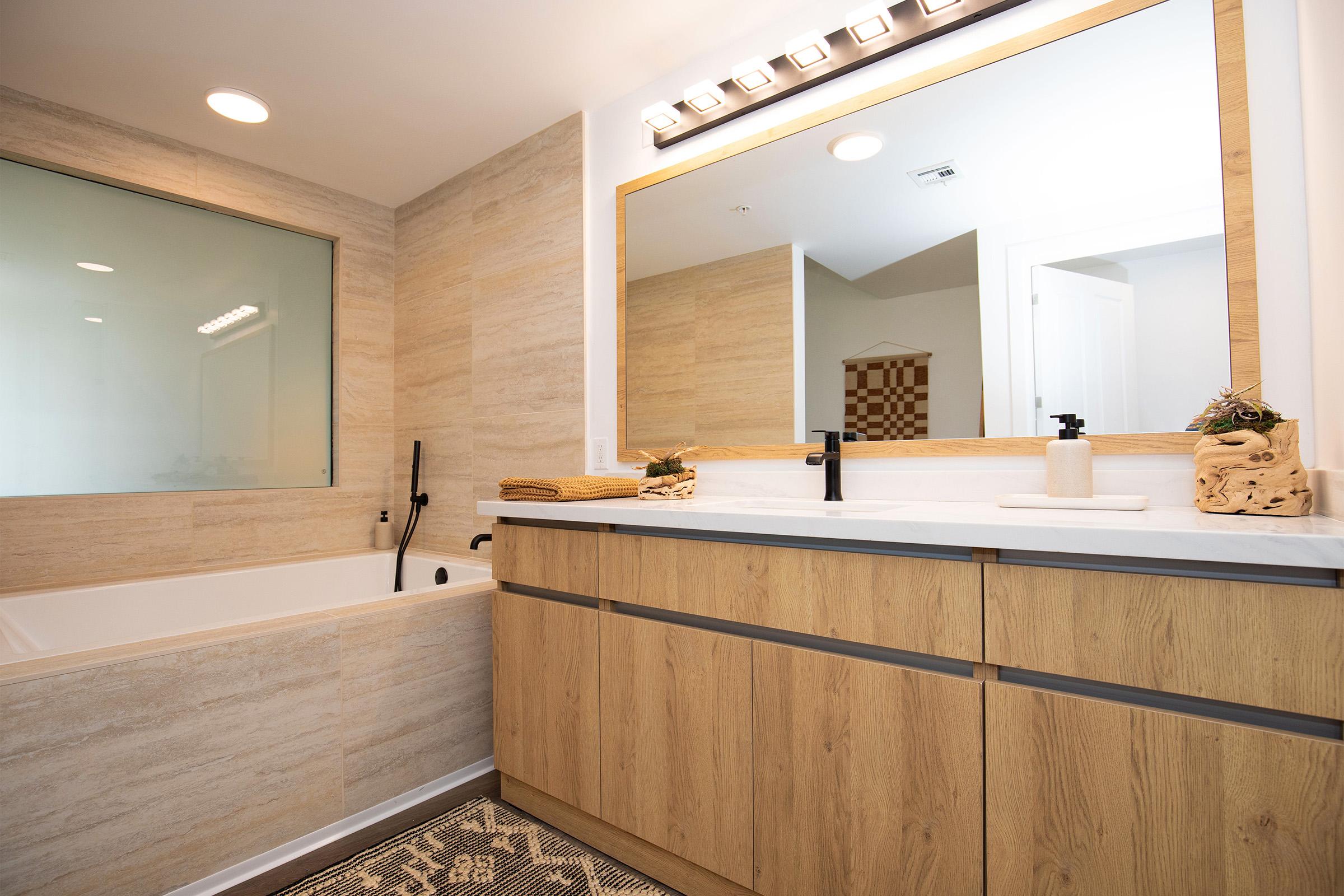 A modern bathroom featuring a light wood vanity with two sinks, a large mirror with warm lighting, a glass shower enclosure, a white bathtub, and neutral-toned walls and flooring. Decorative items include towels and a small plant accent on the counter.