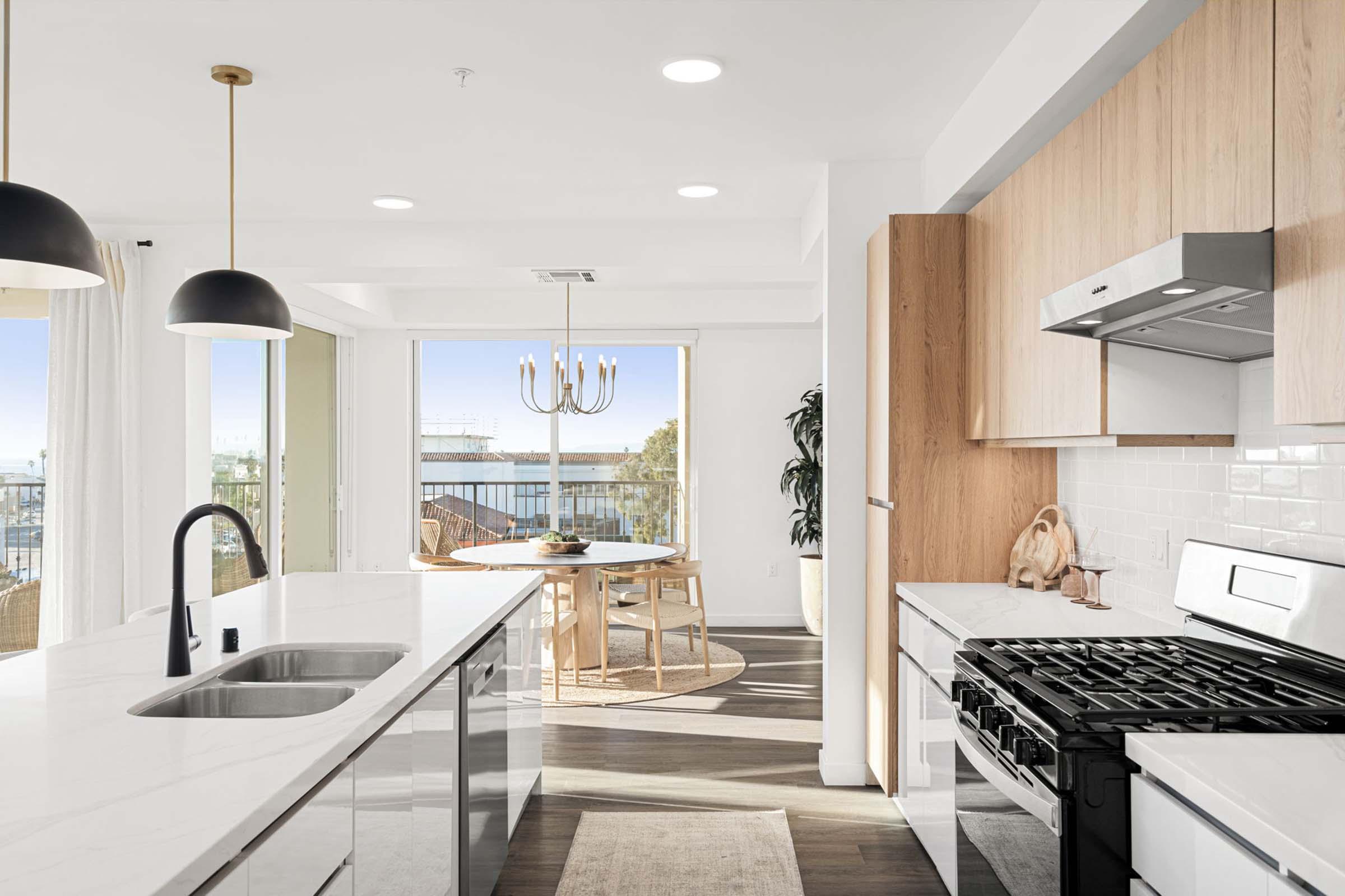 Modern kitchen featuring a large island with a sink, stainless steel appliances, and sleek cabinetry. Natural light streams through large windows, illuminating a dining area with a round table and chairs. The decor blends wood accents with contemporary fixtures, creating a warm and inviting atmosphere.